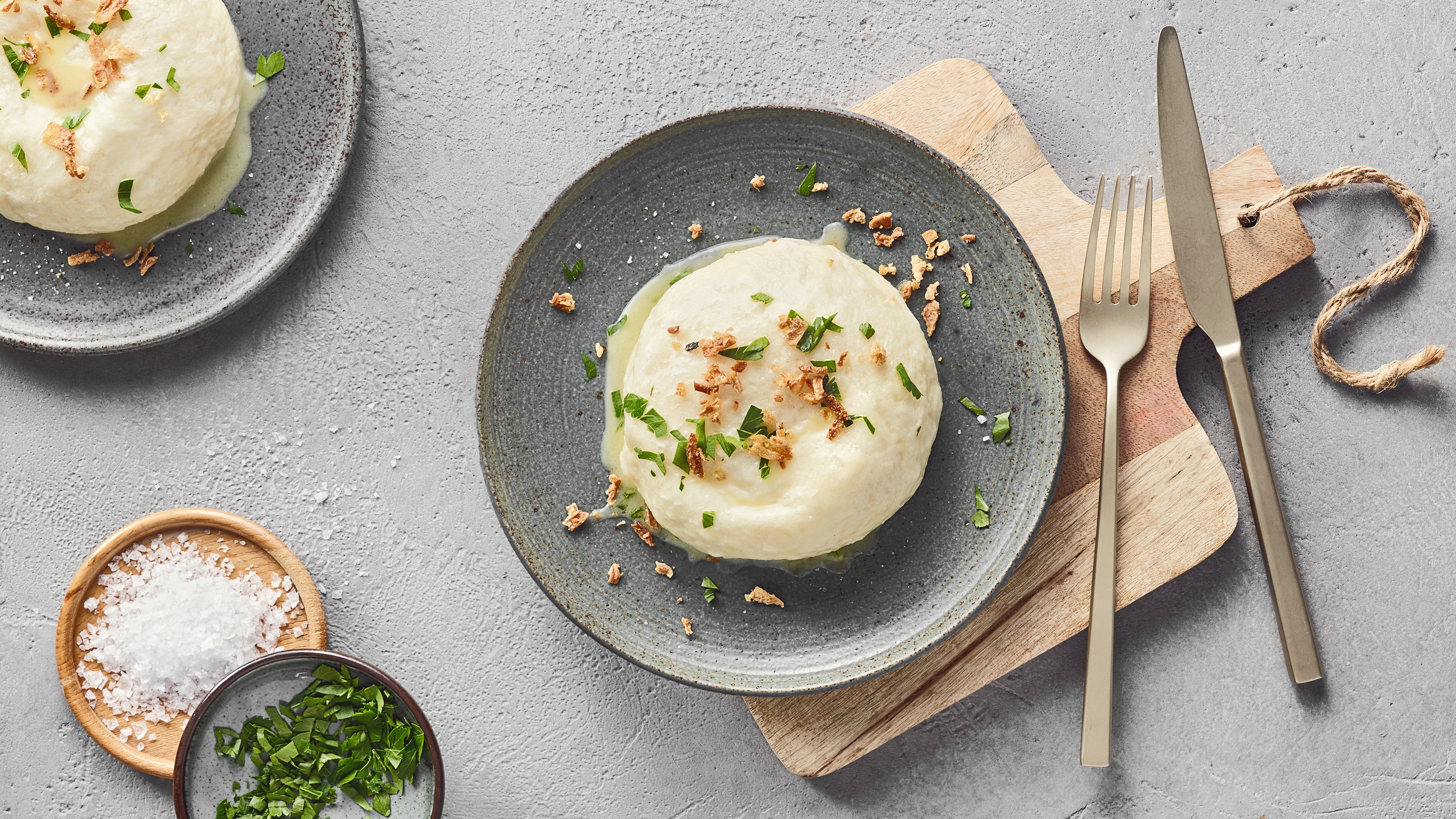 Ein Teller mit einer runden weißen Dampfnudel, darauf grüne Kräuter und braune Krümel.