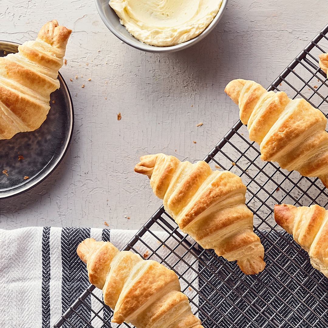 Knusprige, goldgelb gebackene Croissants auf einem Abkühlgitter, daneben ein Schälchen mit luftiger Butter