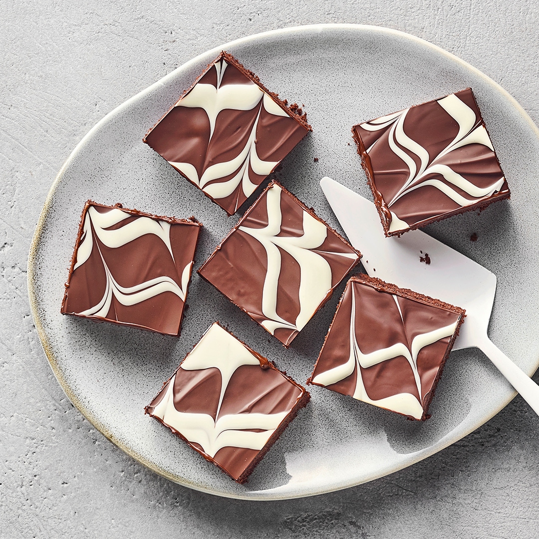 Grauer Teller mit sechs quadratischen Schokoladenkuchenstücken, verziert mit einer sahnigen Garnitur. Dazwischen liegt ein Kuchenheber.