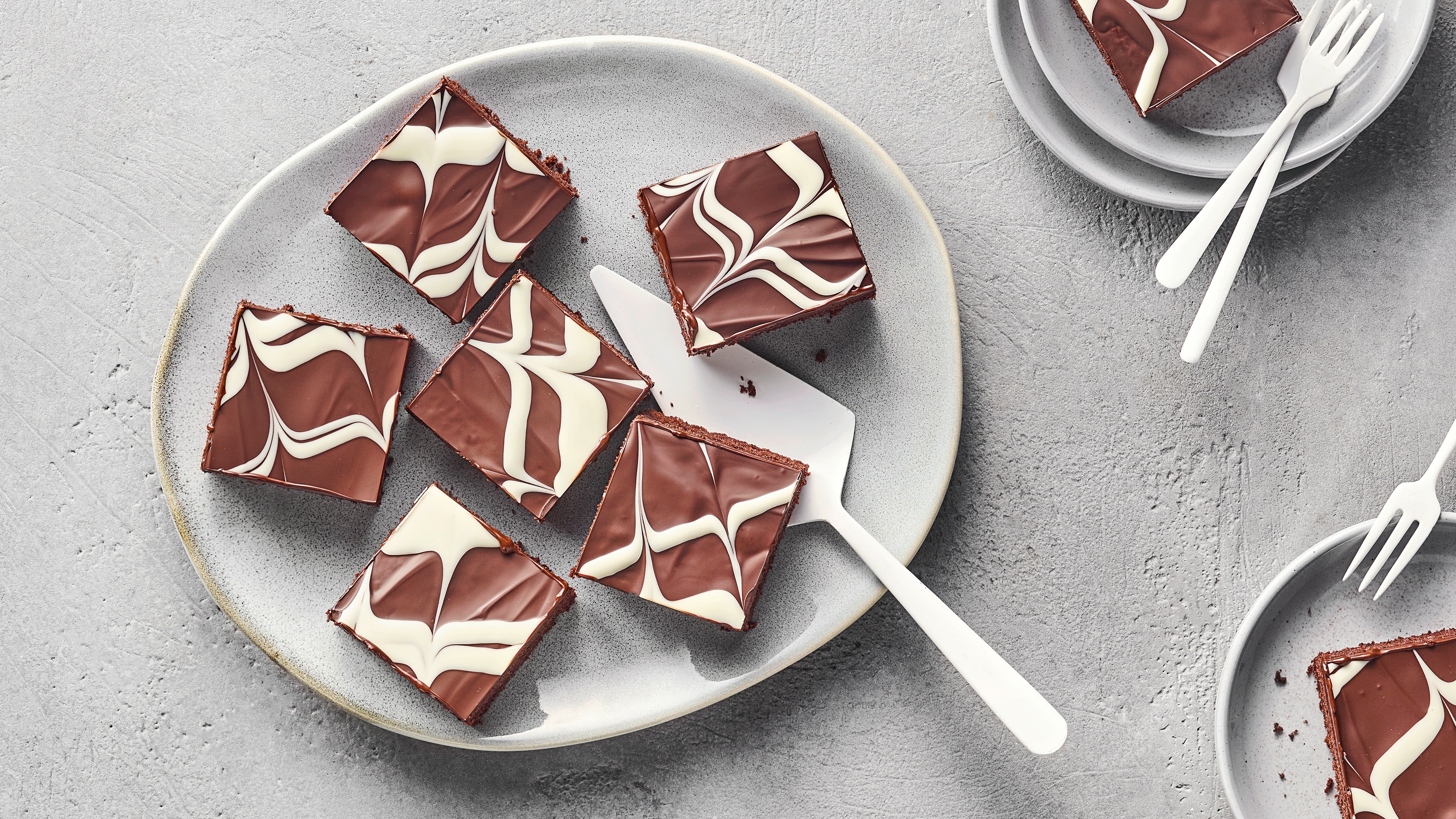 Grauer Teller mit sechs quadratischen Schokoladenkuchenstücken, verziert mit einer sahnigen Garnitur. Dazwischen liegt ein Kuchenheber. Oben rechts und oben links stehen zwei weitere Teller, auf denen jeweils ein einzelnes Stück Kuchen liegt.
