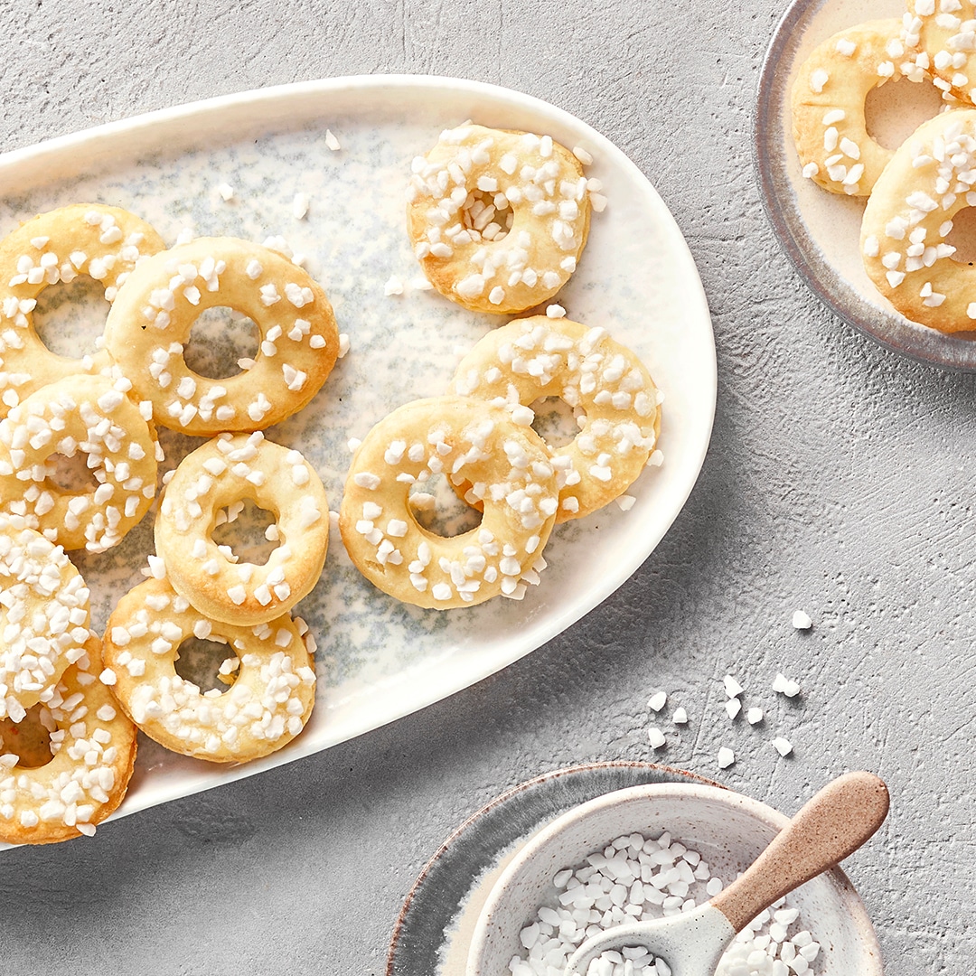 Kleine Ringkekse mit Zuckerguss und Hagelzucker auf einer weißen Servierplatte, daneben eine Schale mit Zucker.