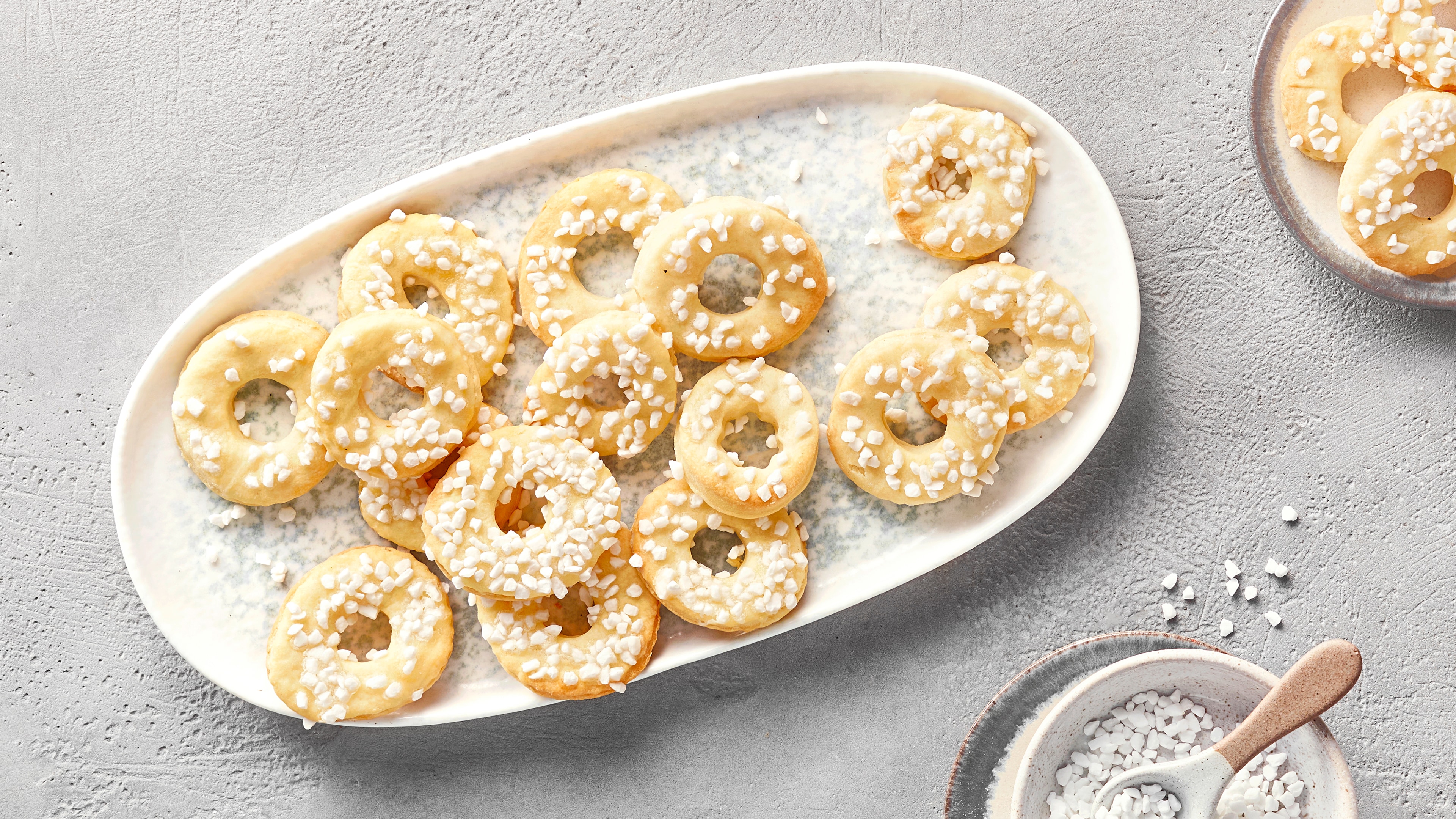 Kleine Ringkekse mit Zuckerguss und Hagelzucker auf einer weißen Servierplatte, daneben eine Schale mit Zucker.