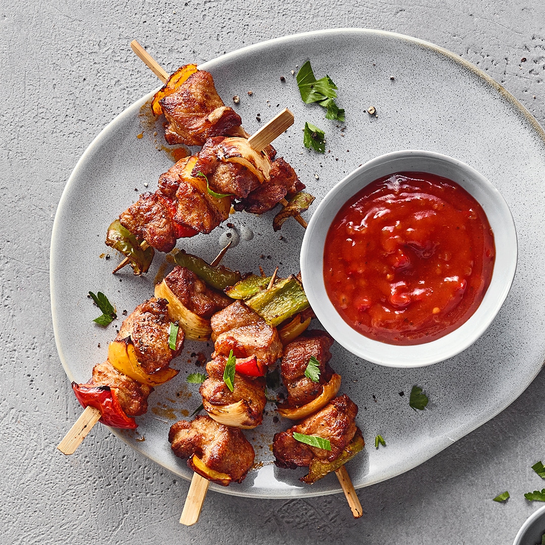 Ein Teller mit gegrillten Fleischspießen, roten, gelben und grünen Paprikastücken, Zwiebelstücken und einer Schale roter Soße.