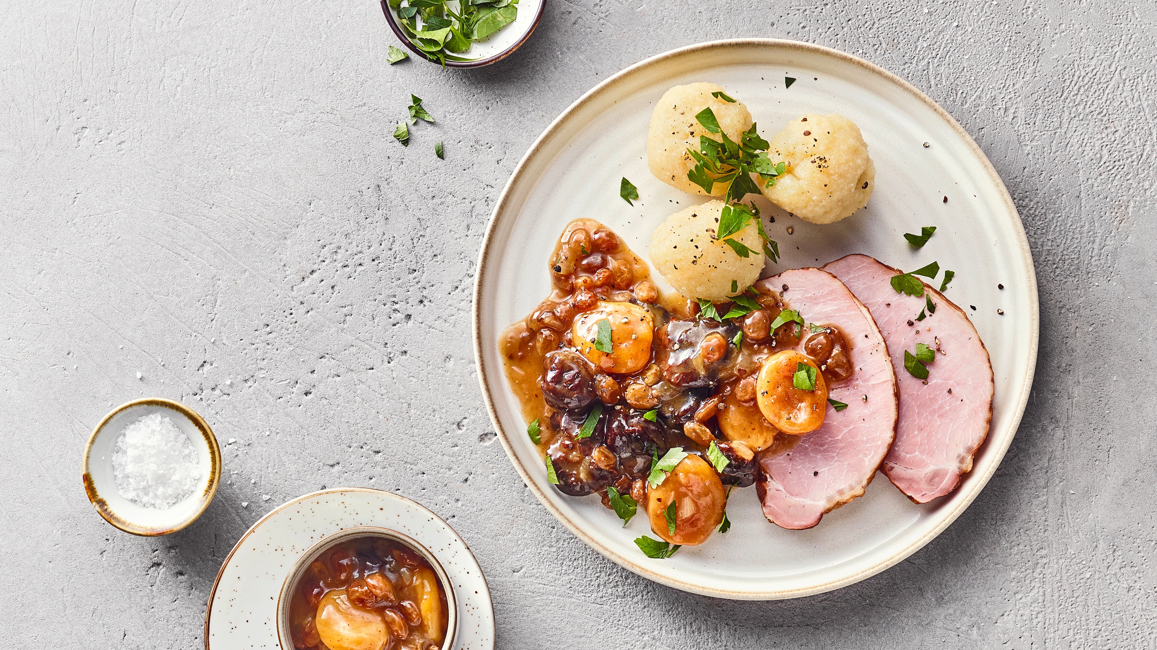 Fleischgericht bestehend aus geräuchertem Schweinebauch, Backobst und Semmelknödeln, verfeinert mit Petersilie. Links daneben stehen Schüsseln mit mehr Backobst, Petersilie und Salz.