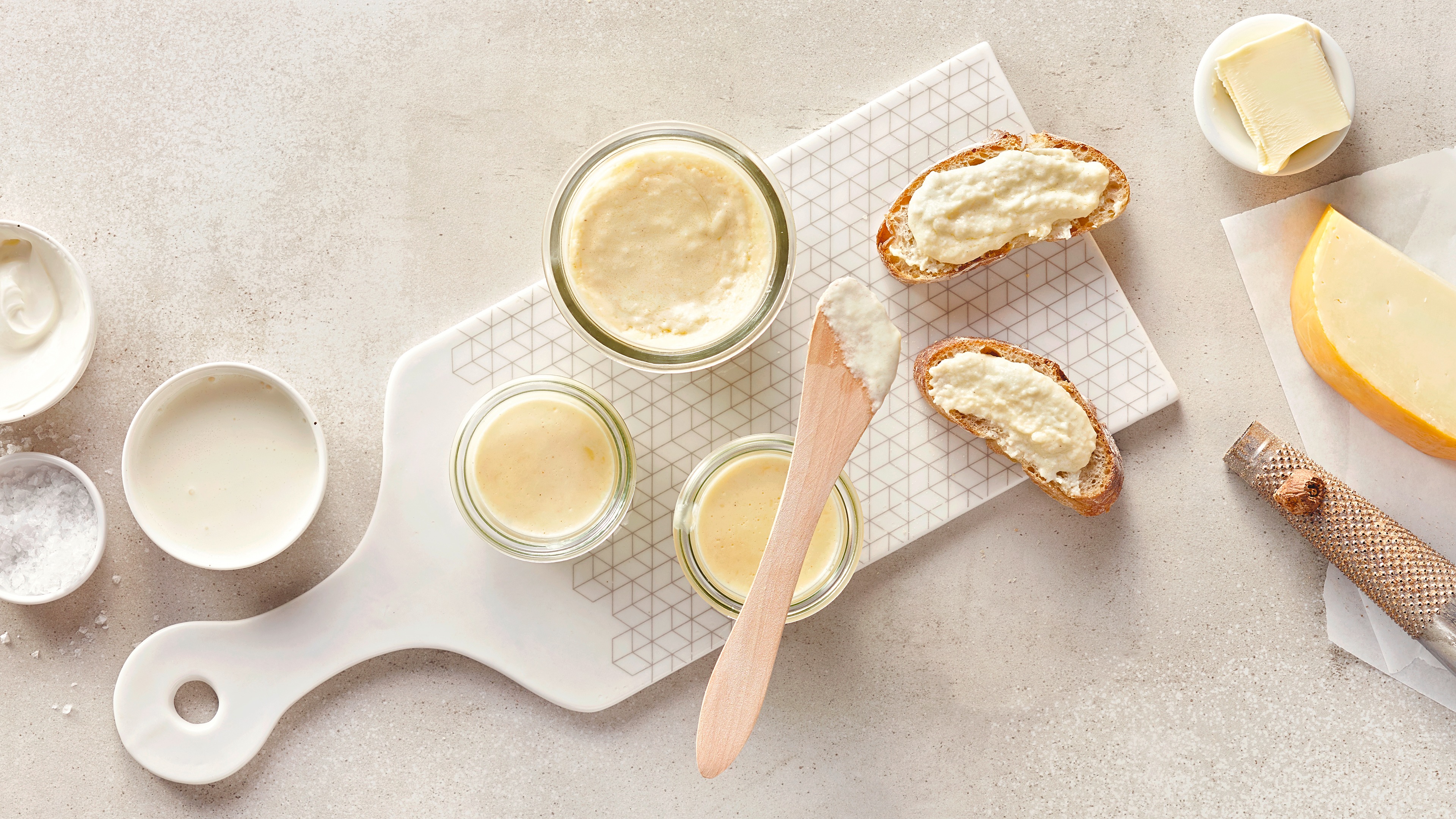 Kleine Glasschüsseln auf einem Brett, gefüllt mit heller Creme, daneben zwei Stücke Brot, bestrichen mit der hellen Creme