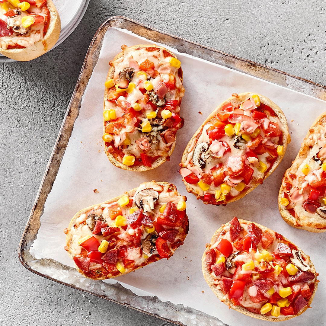 Mehrere überbackene Pizzabrote mit Käse, Paprika, Mais und Champignons auf einem Backblech.