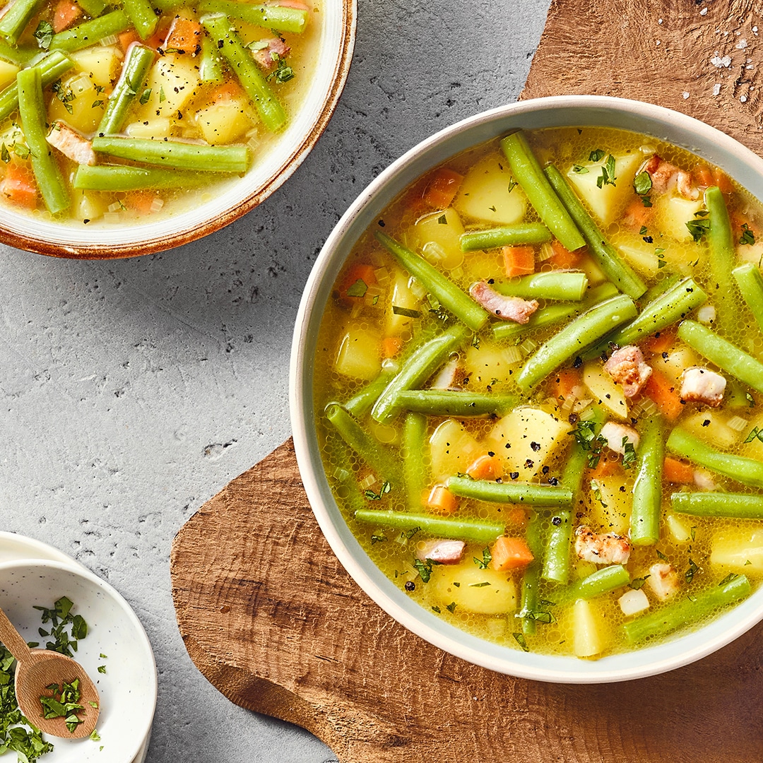 Eine Schüssel mit grünen Bohnen, Kartoffelstücken, Möhrenstücken, kleinen Speckwürfeln und Kräutern in gelber Brühe.