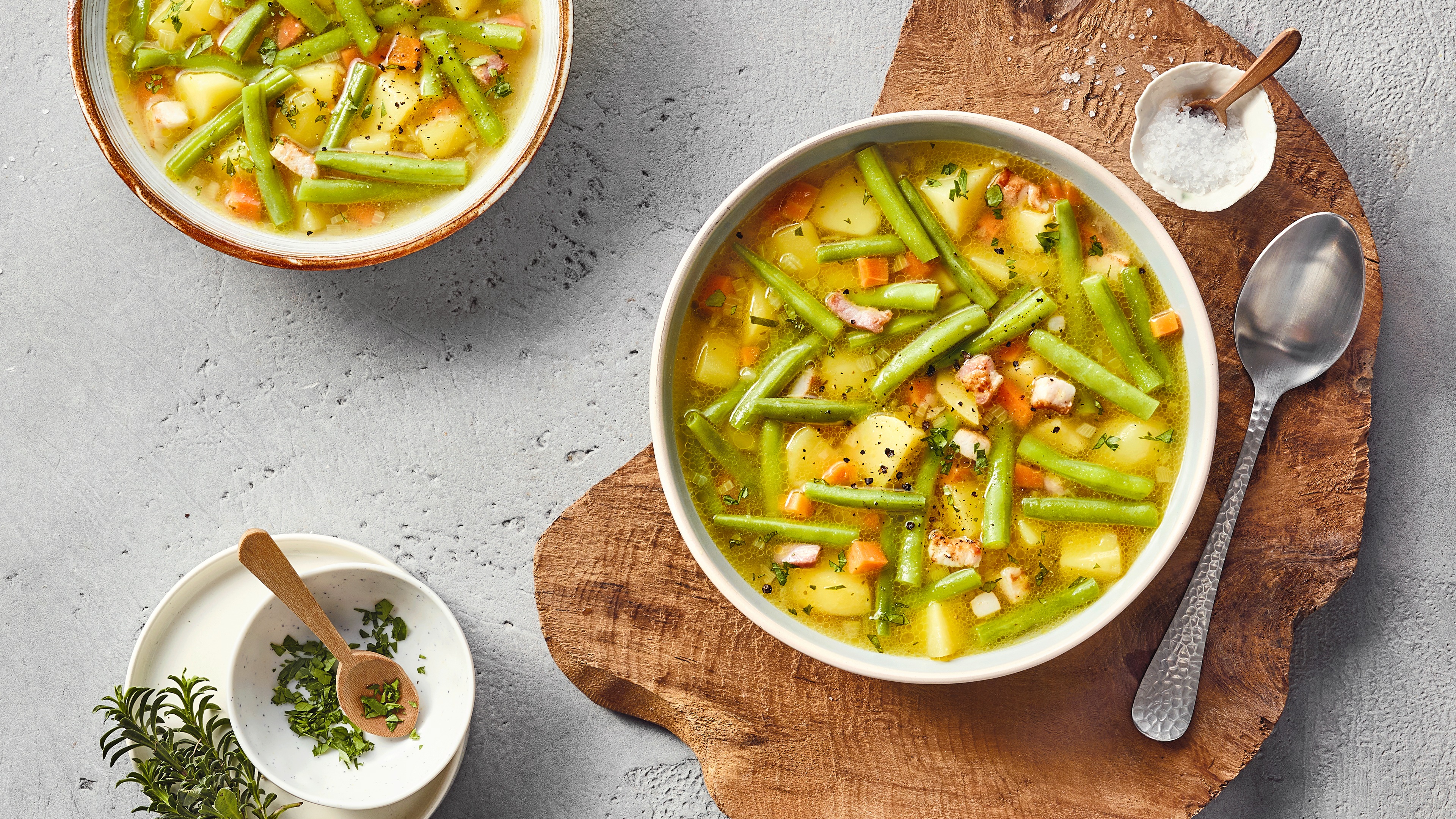 Eine Schüssel mit grünen Bohnen, Kartoffelstücken, Möhrenstücken, kleinen Speckwürfeln und Kräutern in gelber Brühe.