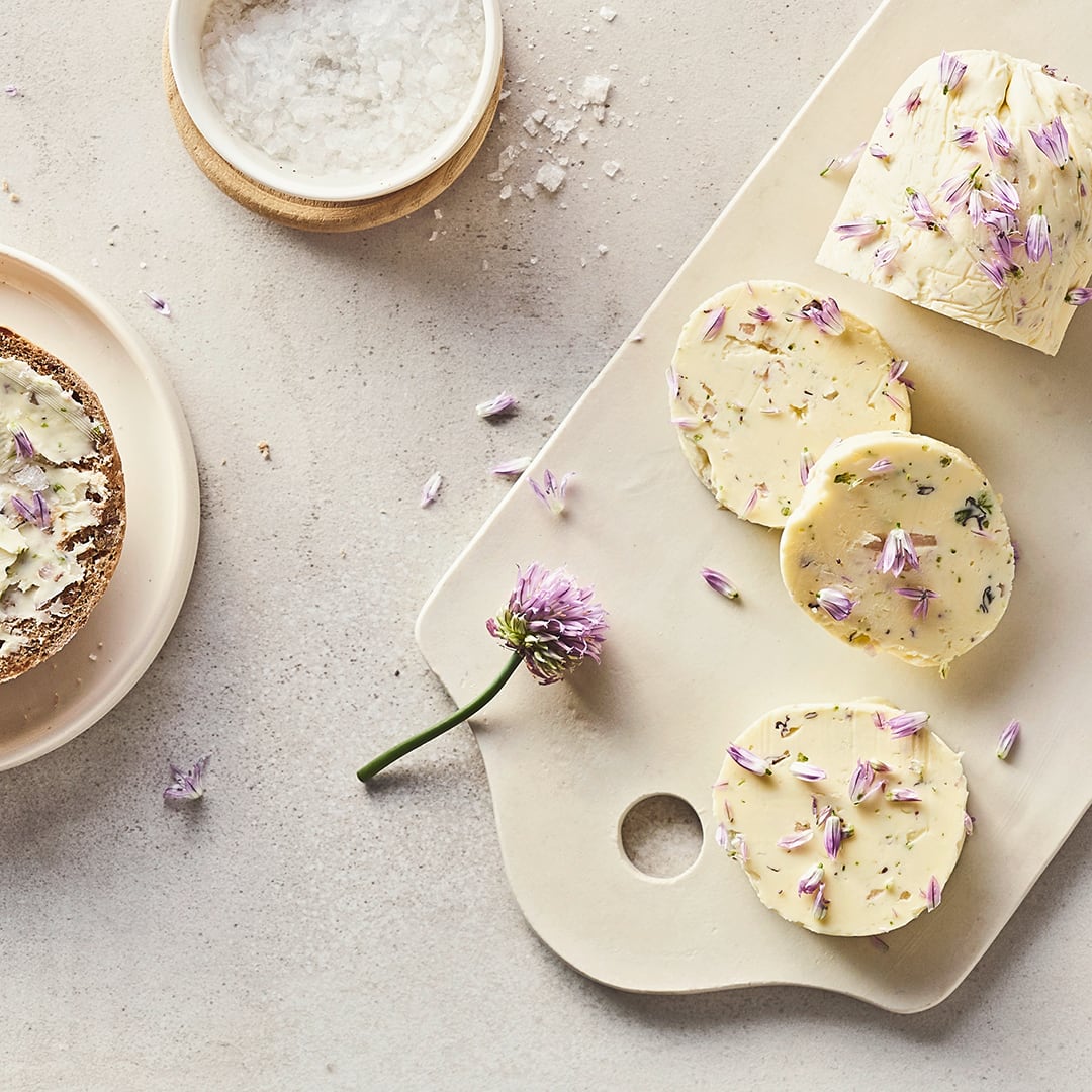 Stückchen Butter mit Schnittlauch, getoppt mit lilafarbenen Blüten, auf einem weißen Brett