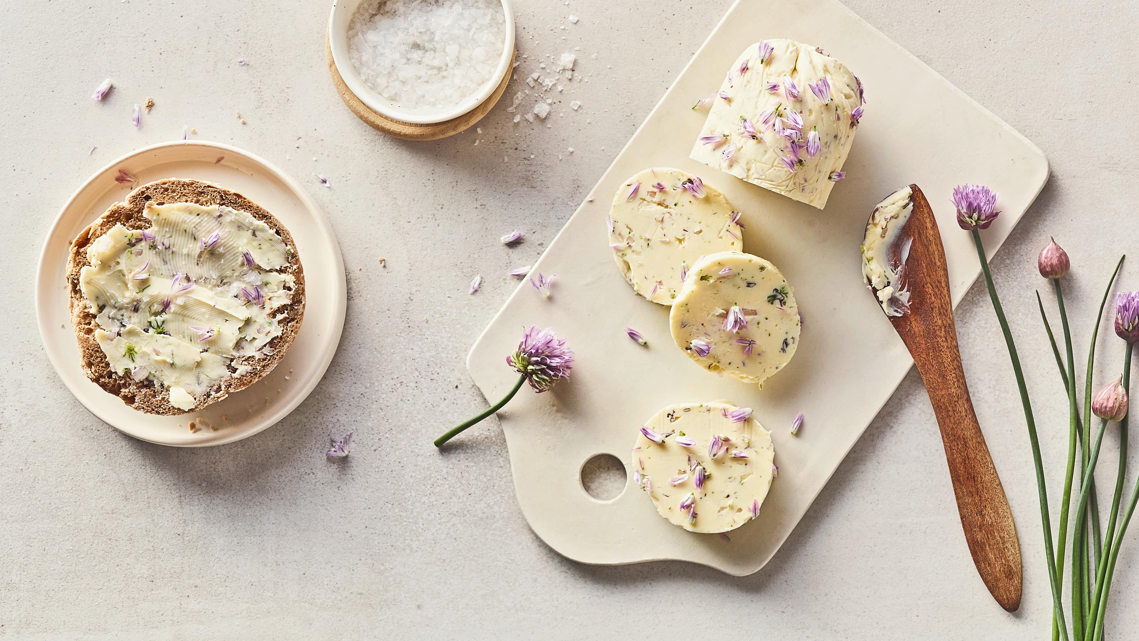 Rezept für Schnittlauchblütenbutter Stückchen Butter mit Schnittlauch, getoppt mit lilafarbenen Blüten, auf einem weißen Brett