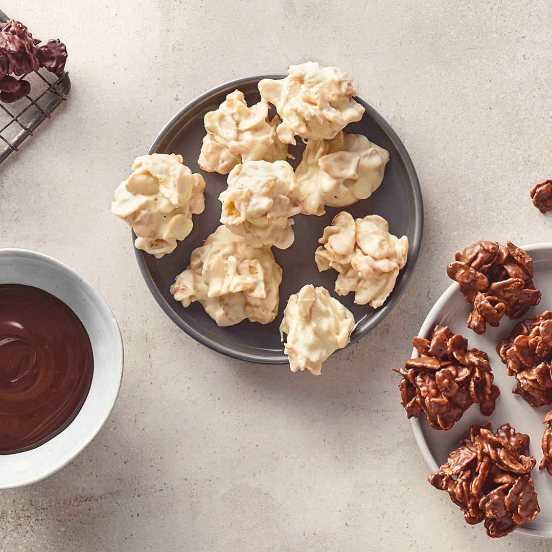 Ein Teller mit weißen und braunen Schoko-Clustern aus knusprigen Zutaten, daneben eine Schale mit flüssiger Schokolade