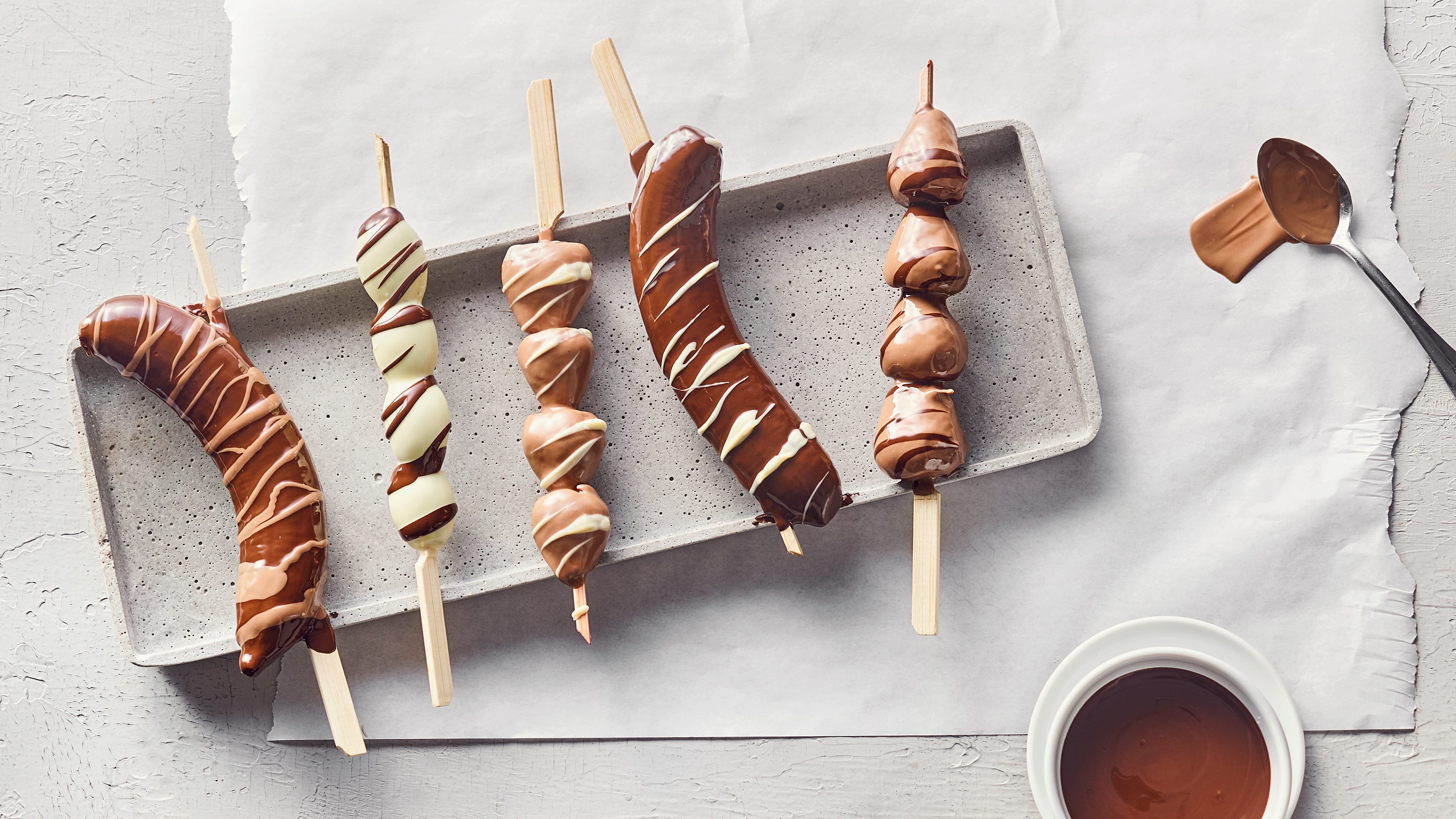 Rezept für Schokofrüchte Ein Steinbrett, dadrauf Erdbeeren und Bananen auf Holzspießen, überzogen mit weißer und dunkler Schokolade