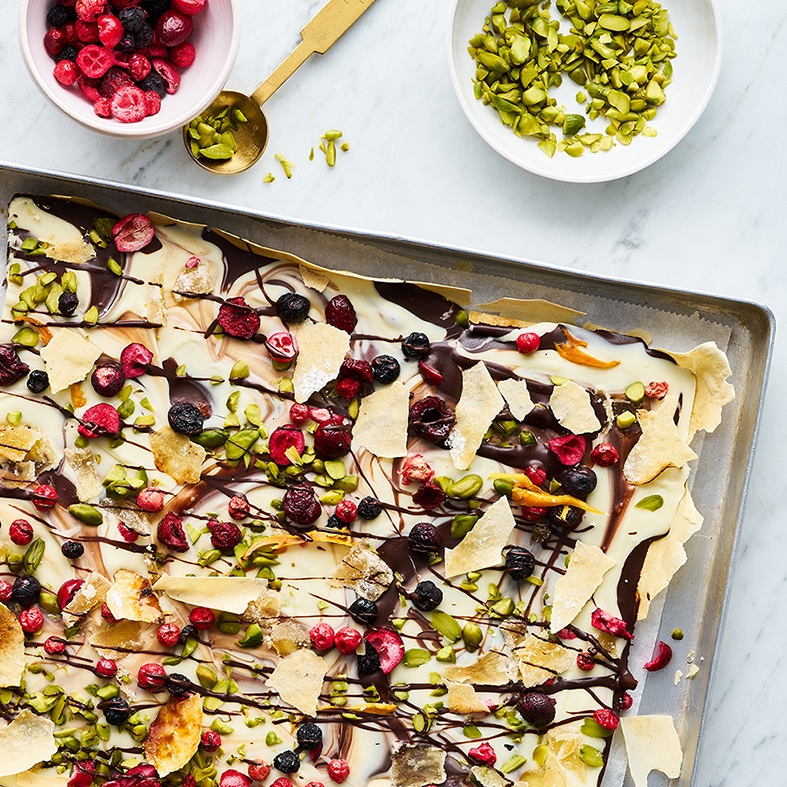 Ein Blech mit Schokoladenbruch, belegt mit getrockneten Beeren, gehackten Pistazien, Schokostreifen und knusprigem Gebäck