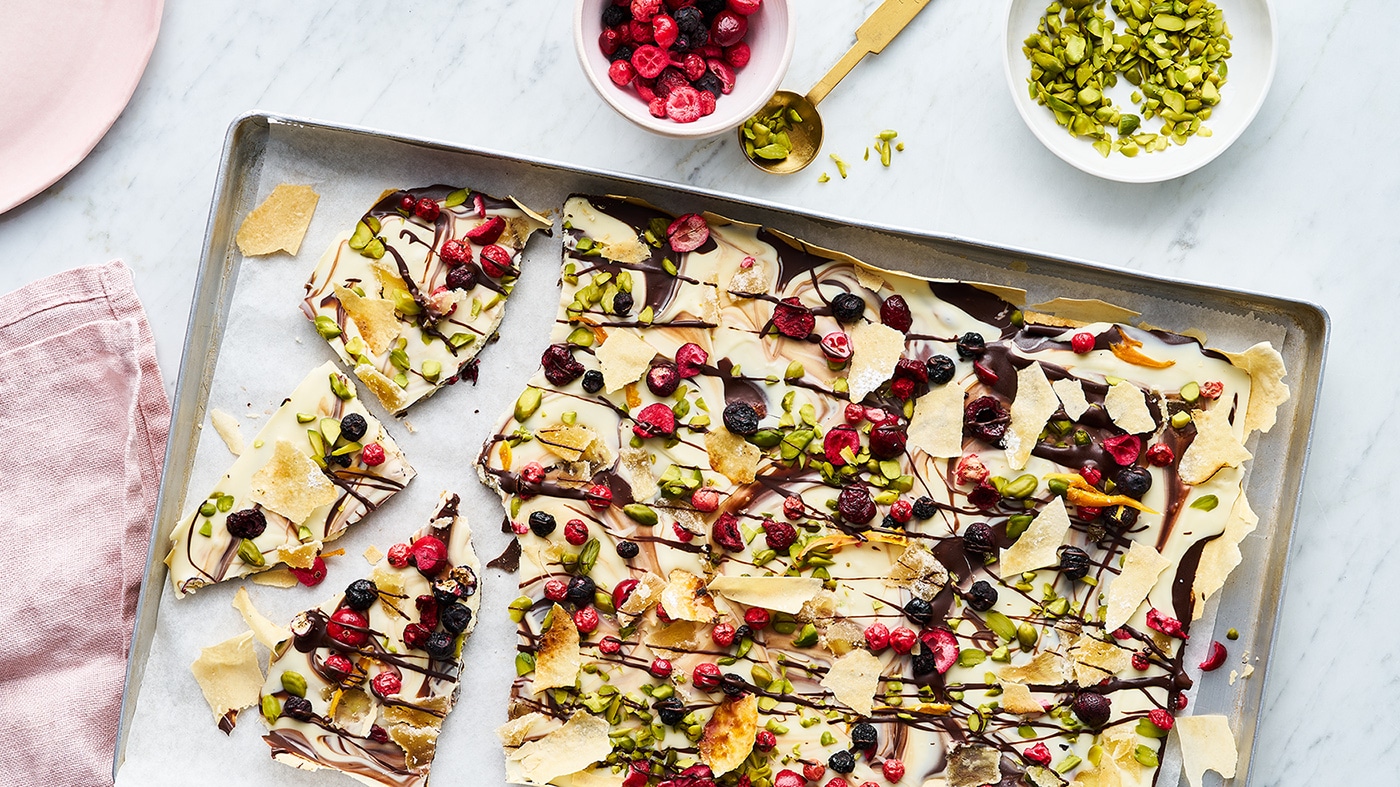 Ein Blech mit Schokoladenbruch, belegt mit getrockneten Beeren, gehackten Pistazien, Schokostreifen und knusprigem Gebäck