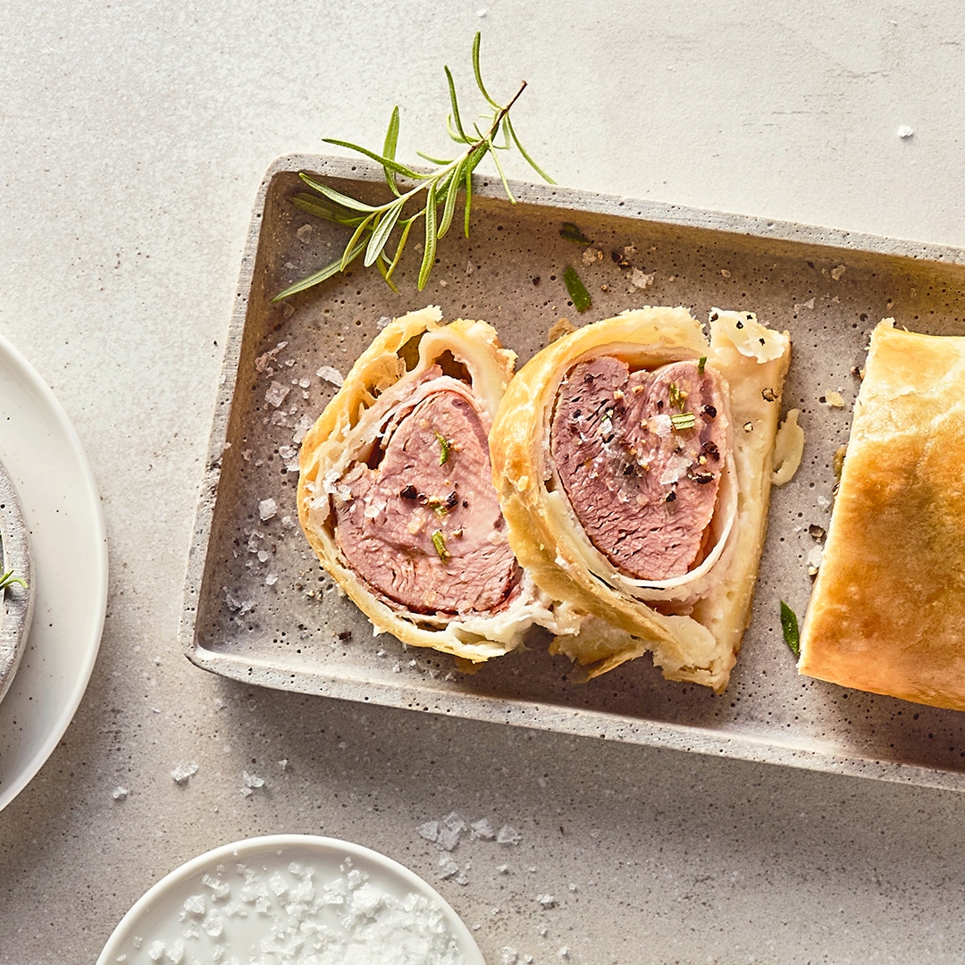 Ein längliches, zartes Stück Fleisch, ummantelt mit gelbgold gebackenem, blättrigem Teig, auf einer Steinplatte