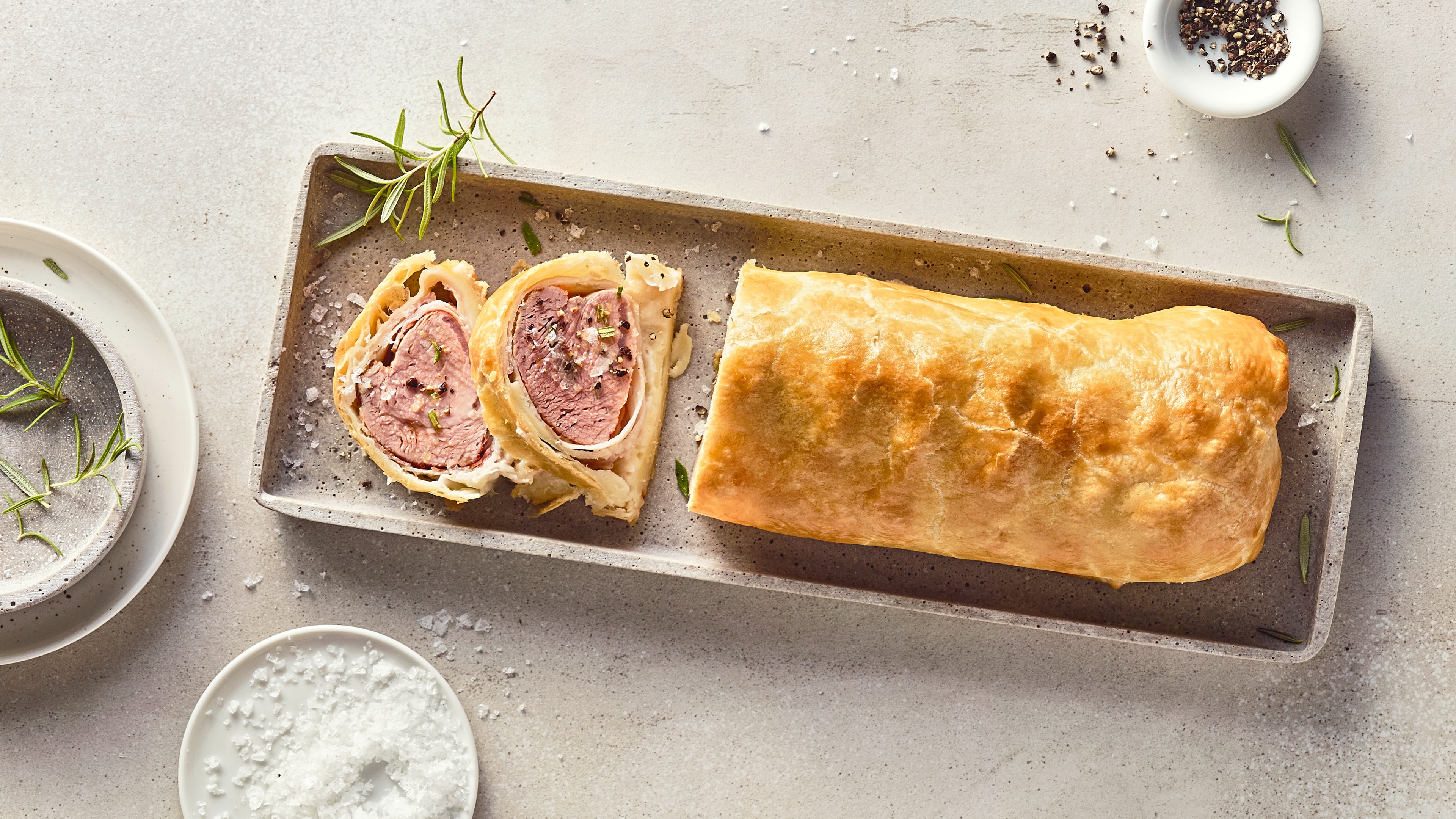 Rezept für Schweinefilet im Blätterteig Ein längliches, zartes Stück Fleisch, ummantelt mit gelbgold gebackenem, blättrigem Teig, auf einer Steinplatte