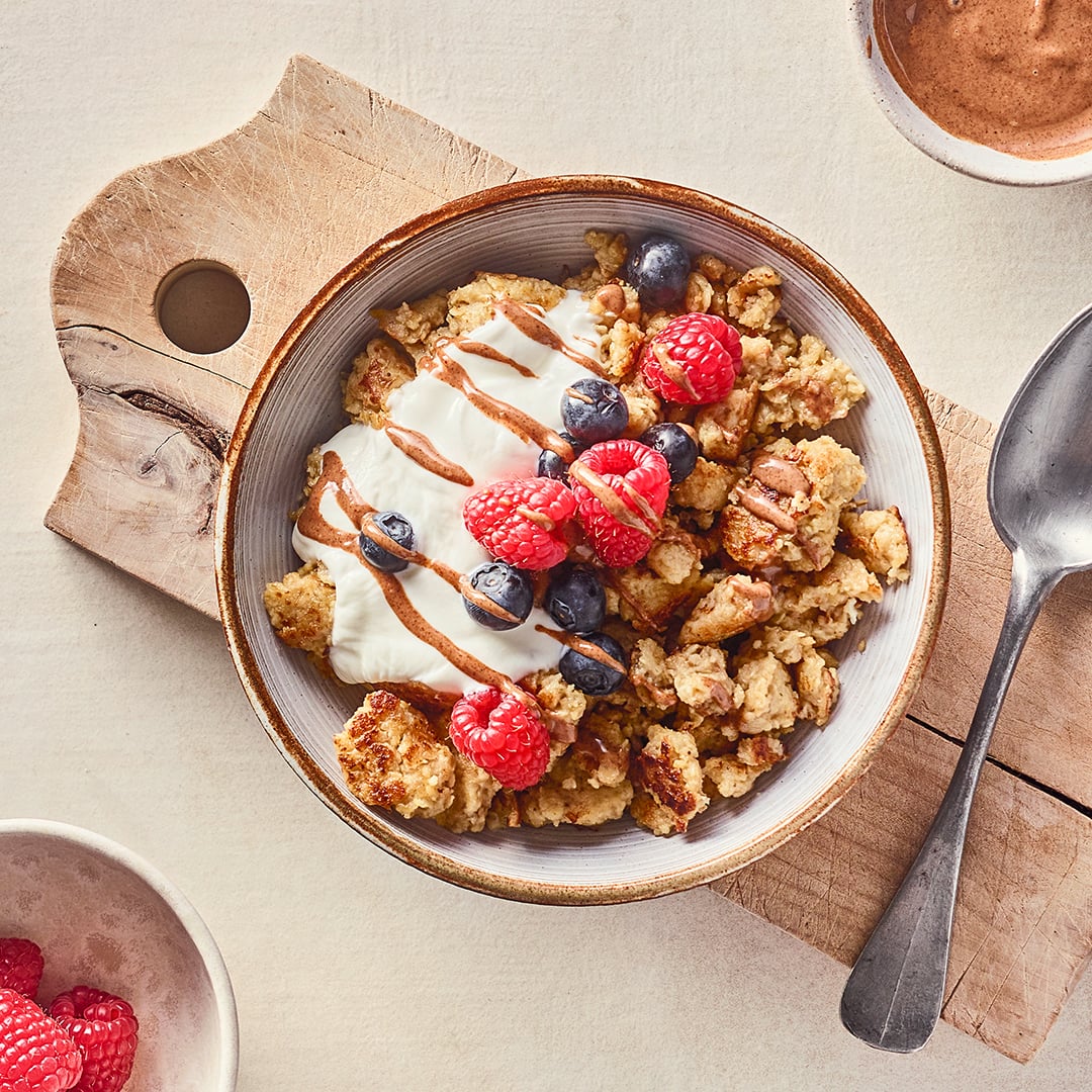 Eine Schale mit Teigstückchen, getoppt mit Joghurt, frischen Blaubeeren, frischen Himbeeren und Nussmuss
