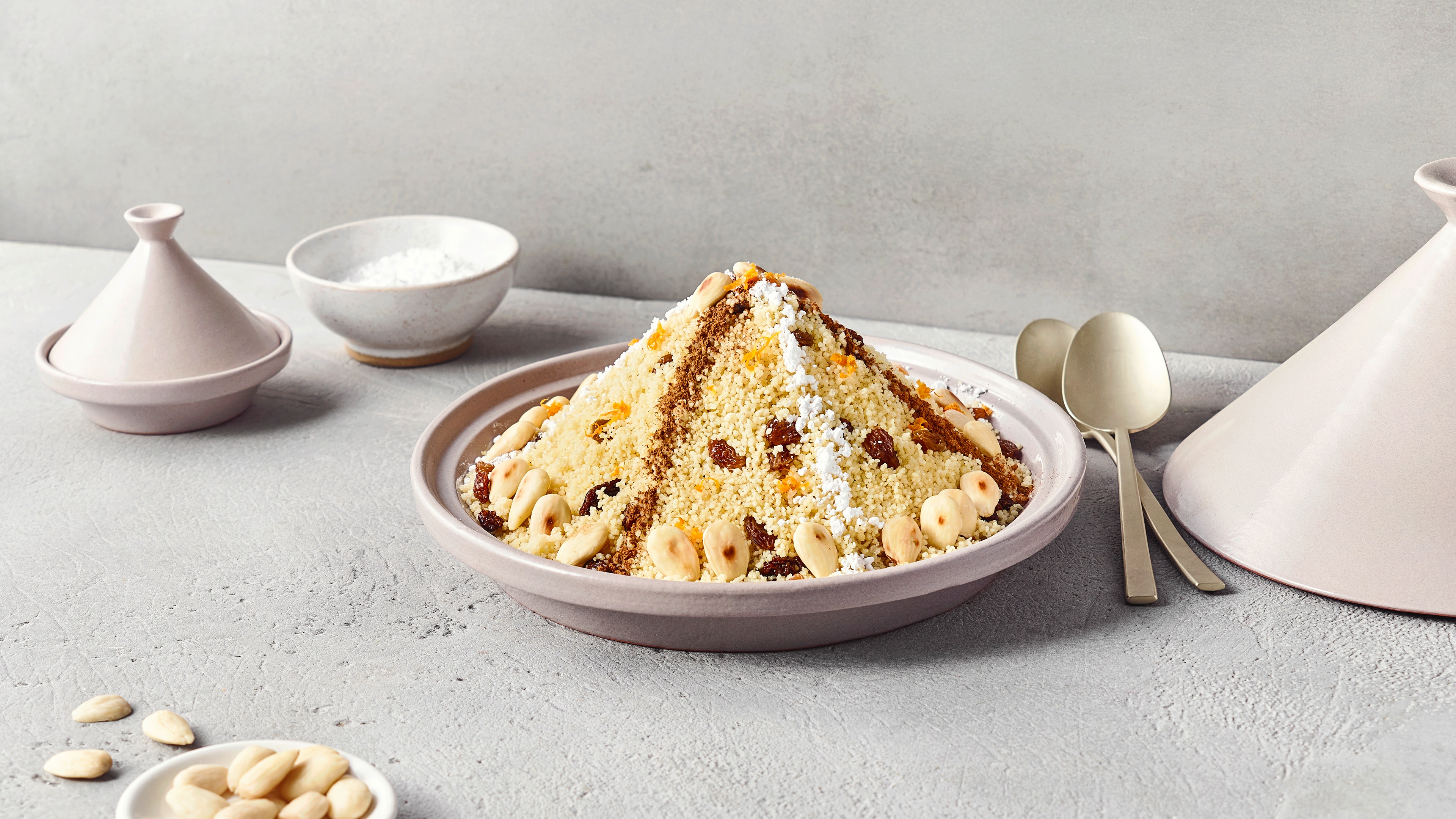 Ein Teller mit gelbem Couscous, weißen Nüssen, braunen Rosinen, weißen Zuckerstreifen und braunen Gewürzstreifen zu einem Hügel geformt.