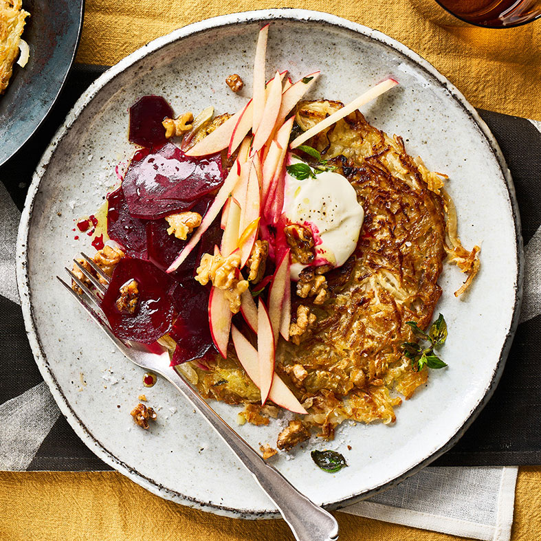 Rösti mit Rote-Bete-Scheiben, Apfelstreifen, Walnüssen und einem Klecks Creme auf einem Teller.