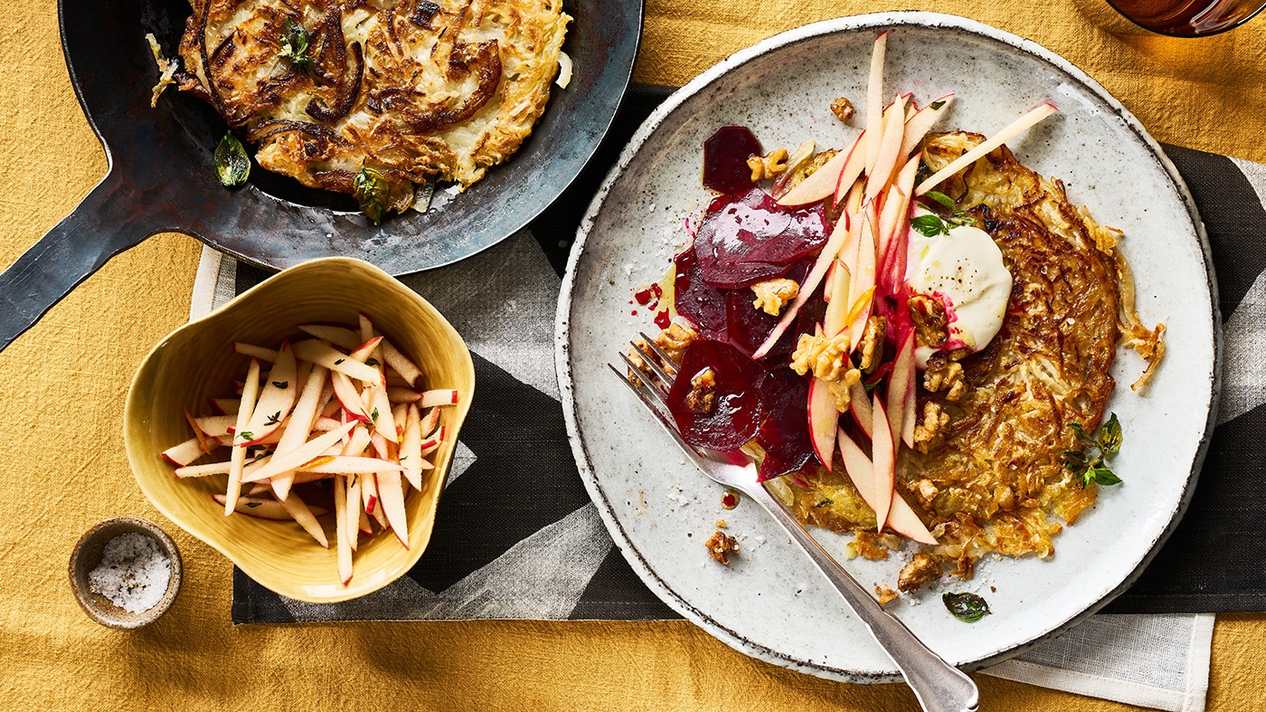 Rösti mit Rote-Bete-Scheiben, Apfelstreifen, Walnüssen und einem Klecks Creme auf einem Teller.