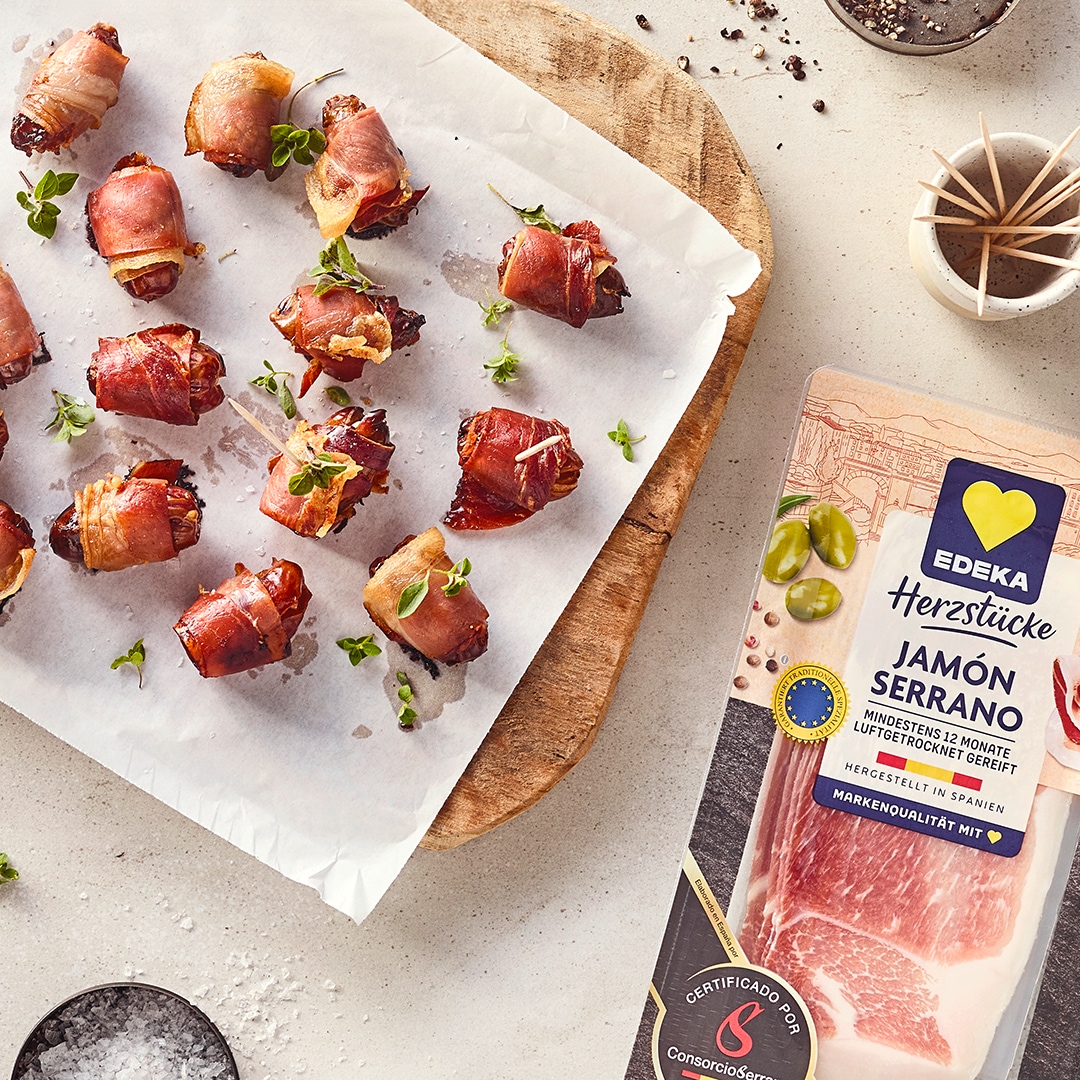 Kleine mit Schinken umwickelte Datteln, fixiert mit Zahnstochern auf hellem Backpapier auf einem Holzbrett mit EDEKA Herzstücke Serrano Schinken Packung