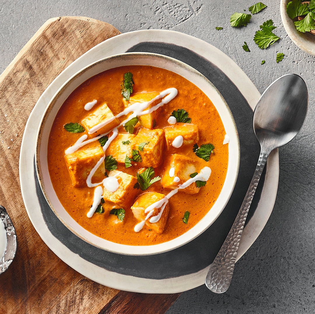 Eine Schale mit orangefarbener Currysoße, darin mehrere Würfel aus Paneer, garniert mit frischen grünen Kräutern und weißen Cremestreifen