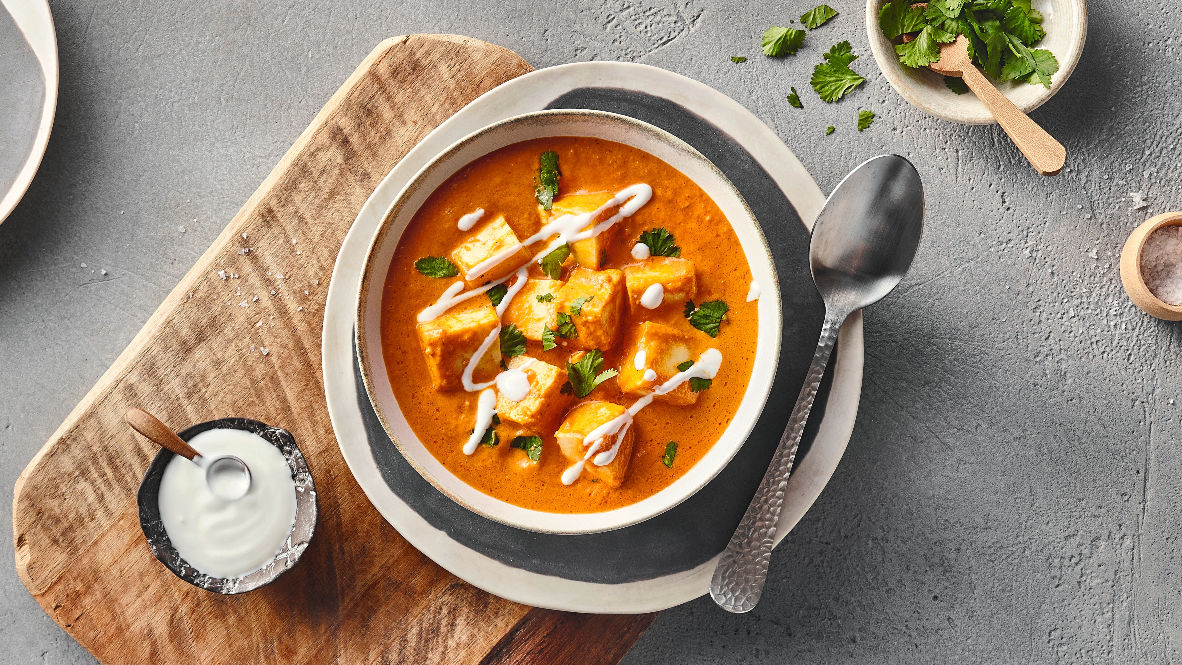 Eine Schale mit orangefarbener Currysoße, darin mehrere Würfel aus Paneer, garniert mit frischen grünen Kräutern und weißen Cremestreifen