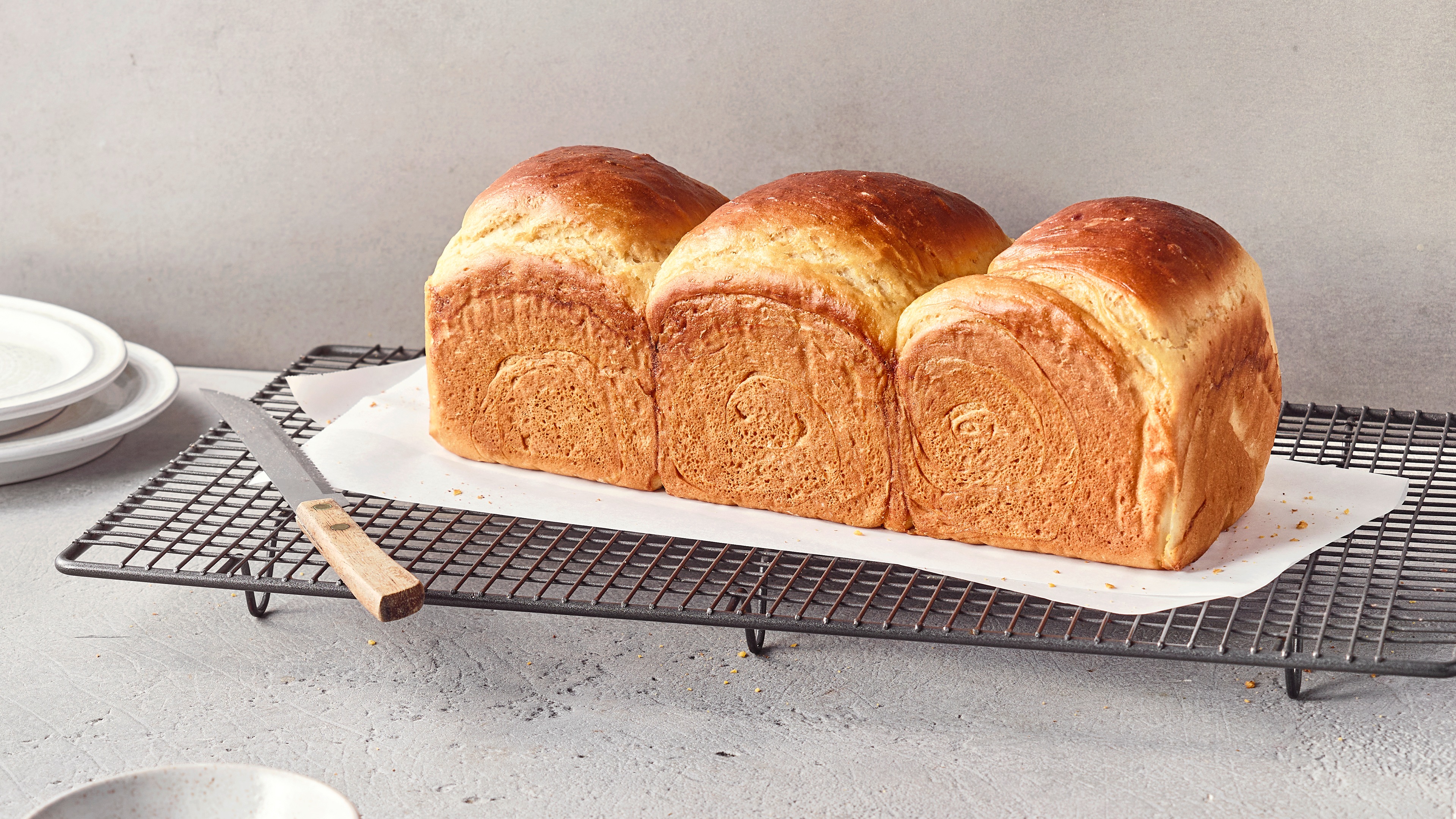 Ein Gitterrost mit drei goldbraunen Brotlaiben mit heller, weicher Kruste auf weißem Backpapier.
