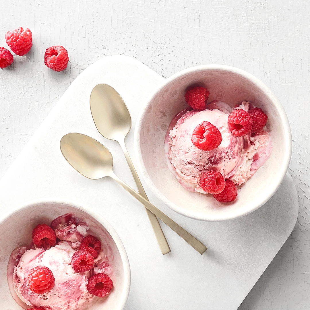 Zwei kleine Schüsseln mit rosafarbener Eiscreme und Himbeeren. In der Mitte liegen zwei Löffel.