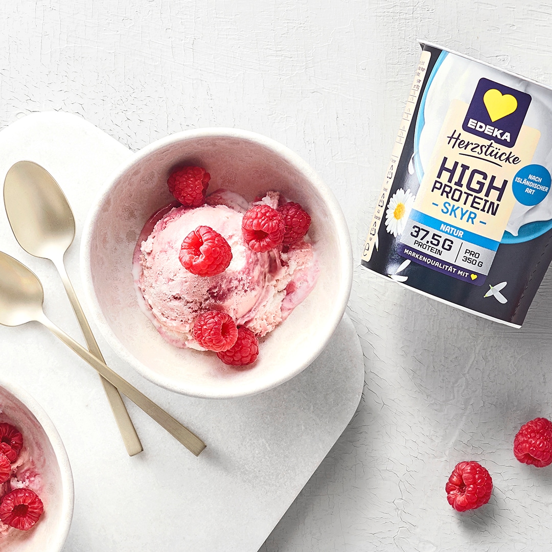 Eine kleine Schüssel mit rosafarbener Eiscreme und Himbeeren. In der Mitte liegen zwei Löffel. Oben rechts liegt das Produkt EDEKA Herzstücke High Protein Skyr