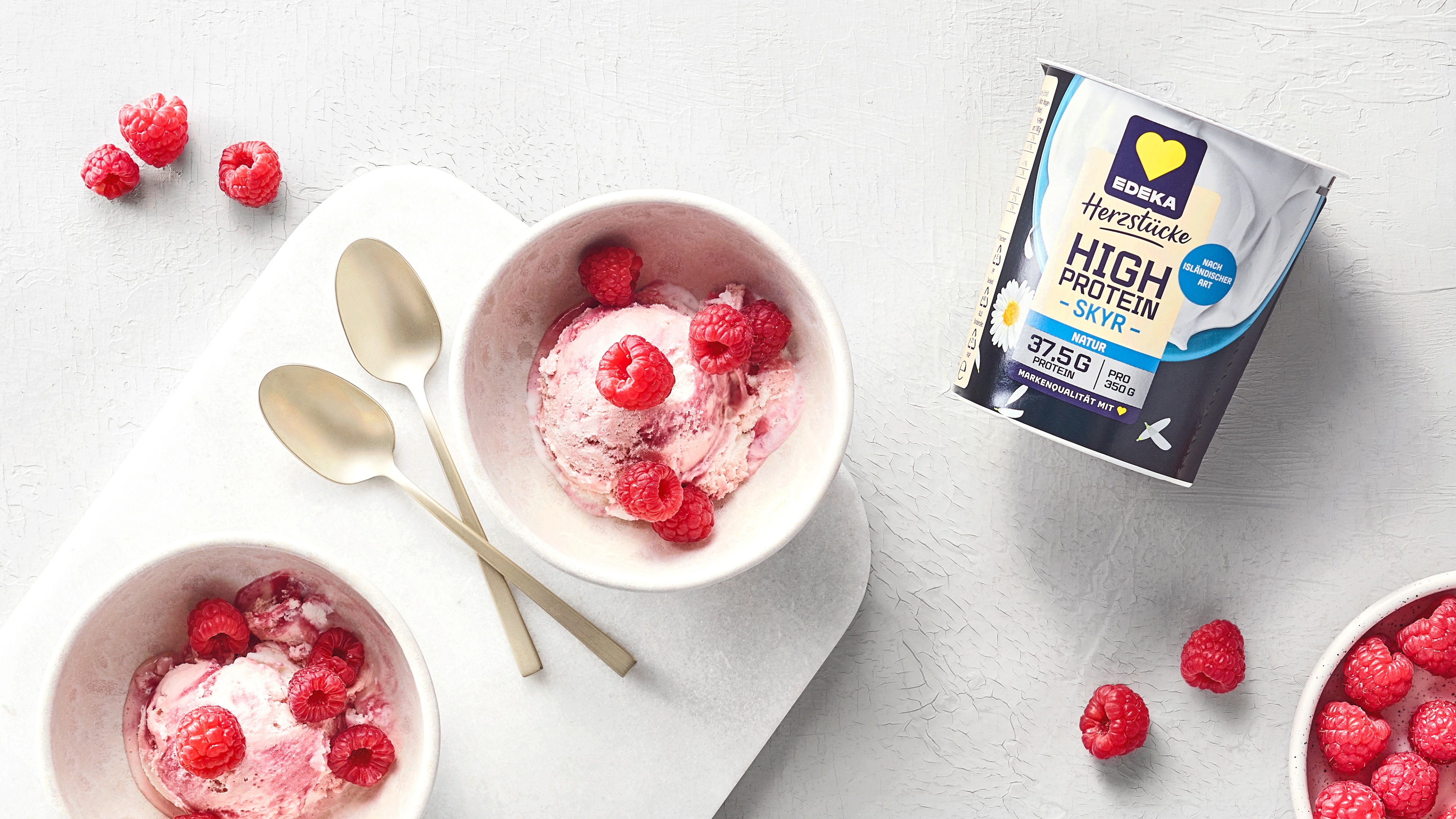 Zwei kleine Schüsseln mit rosafarbener Eiscreme und Himbeeren. In der Mitte liegen zwei Löffel. Oben rechts liegt das Produkt EDEKA Herzstücke High Protein Skyr
