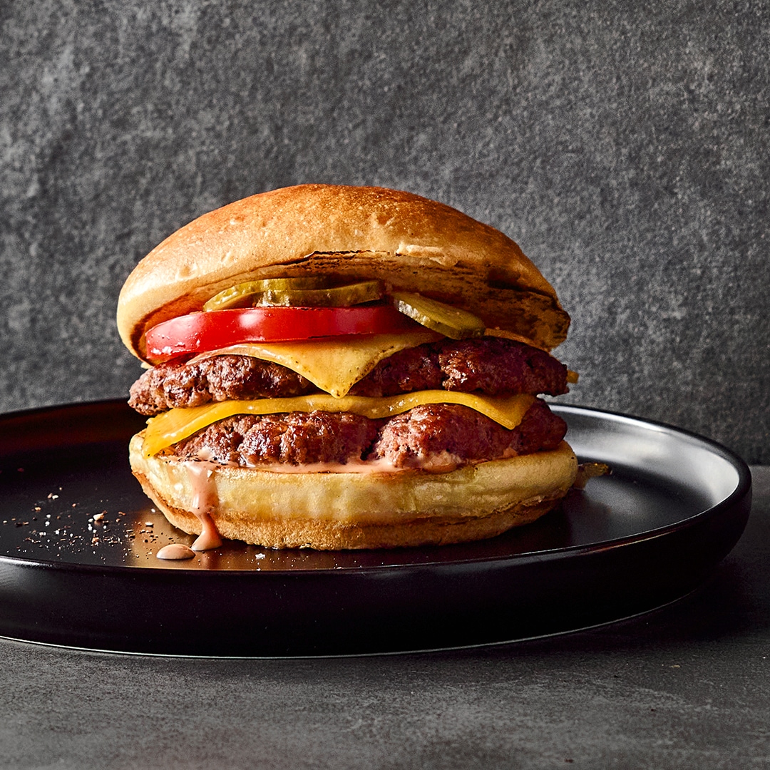 Fluffige Burgerbrötchen mit saftigen Fleischpattys, geschmolzenem Käse, Tomate, Gewürzgurken und hellroter Soße auf runden Tellern