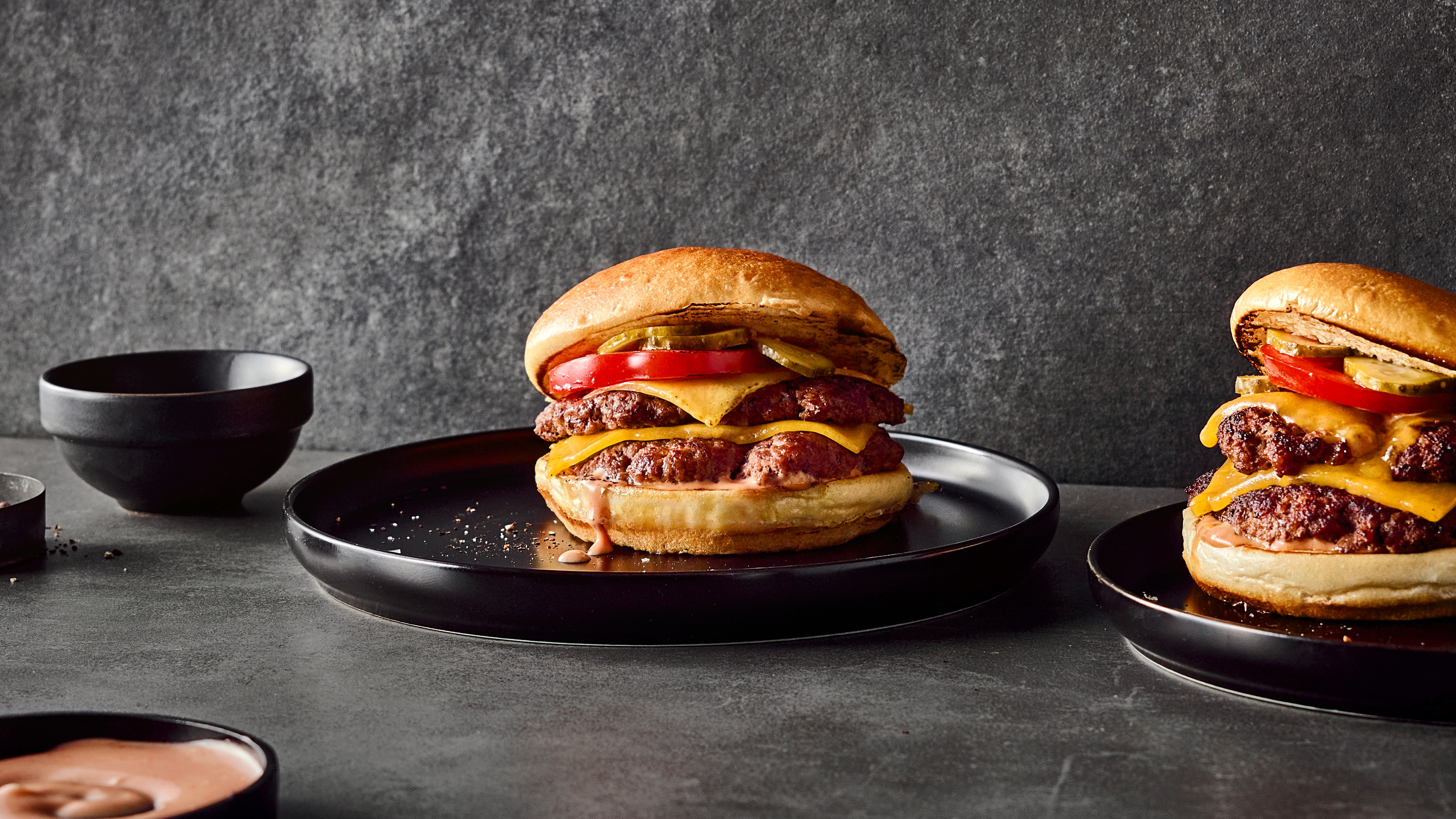 Fluffige Burgerbrötchen mit saftigen Fleischpattys, geschmolzenem Käse, Tomate, Gewürzgurken und hellroter Soße auf runden Tellern