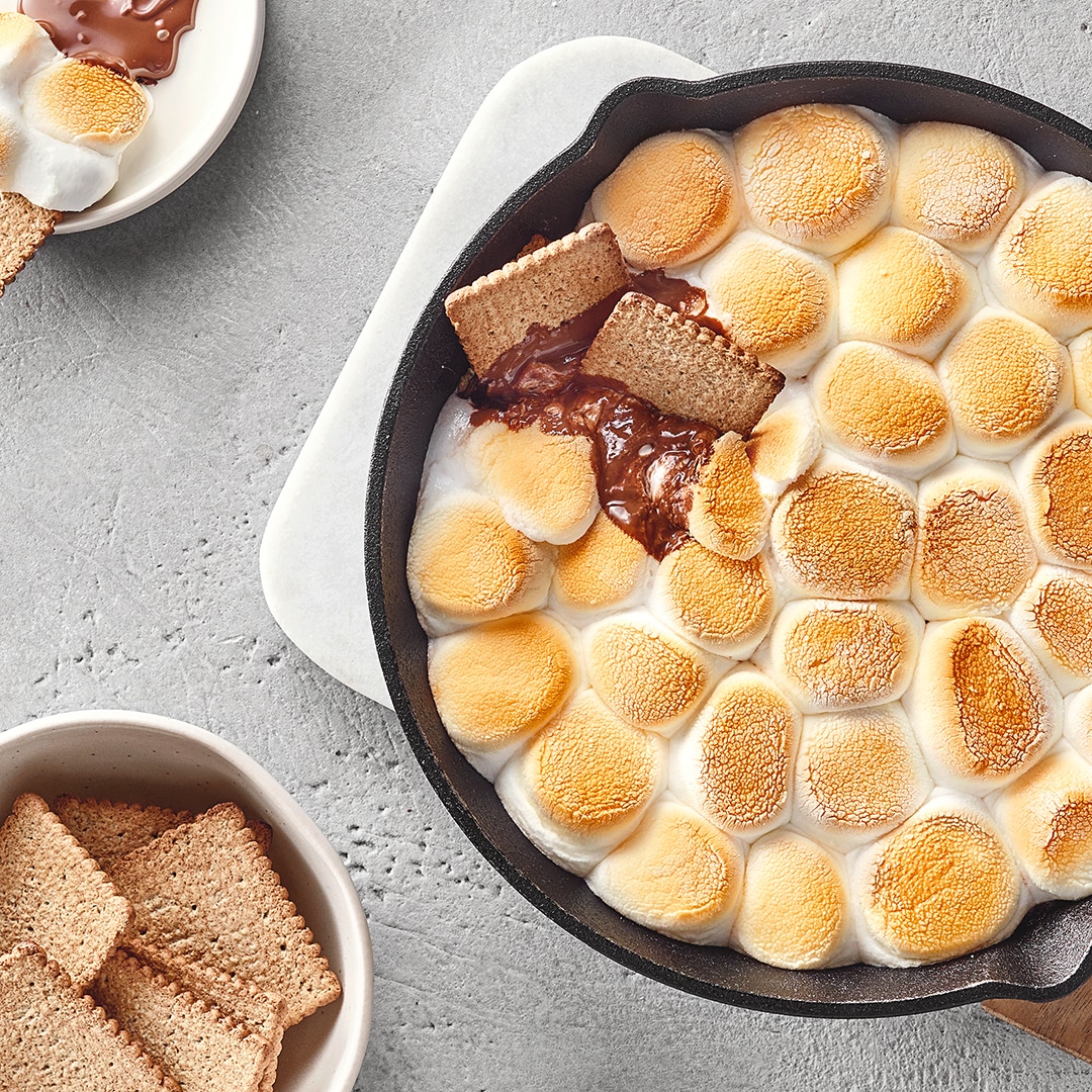 Gusseiserne Pfanne mit gerösteten Marshmallows und geschmolzener Schokolade, serviert mit Keksen auf heller Steinoberfläche.