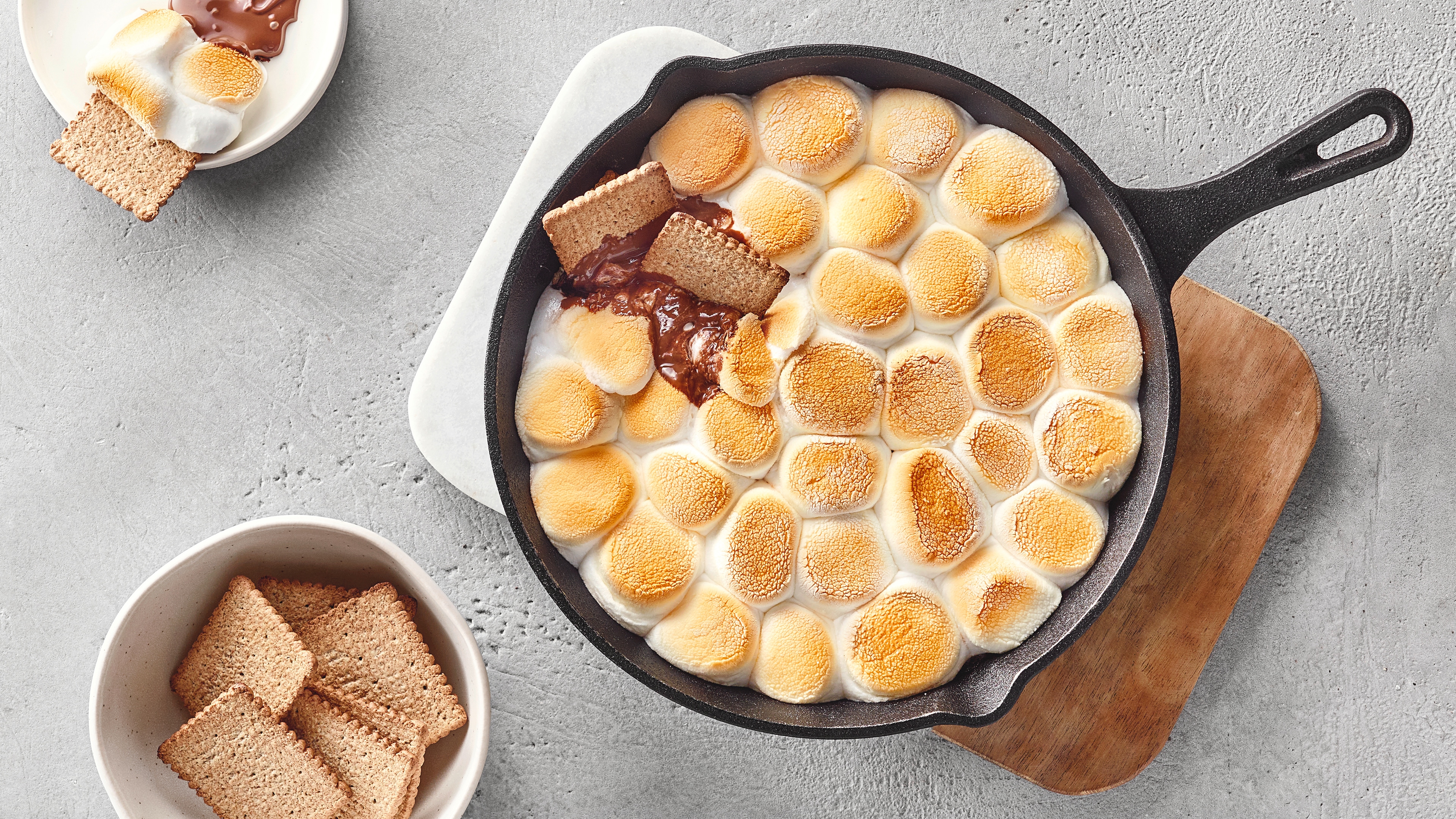 Gusseiserne Pfanne mit gerösteten Marshmallows und geschmolzener Schokolade, serviert mit Keksen auf heller Steinoberfläche.