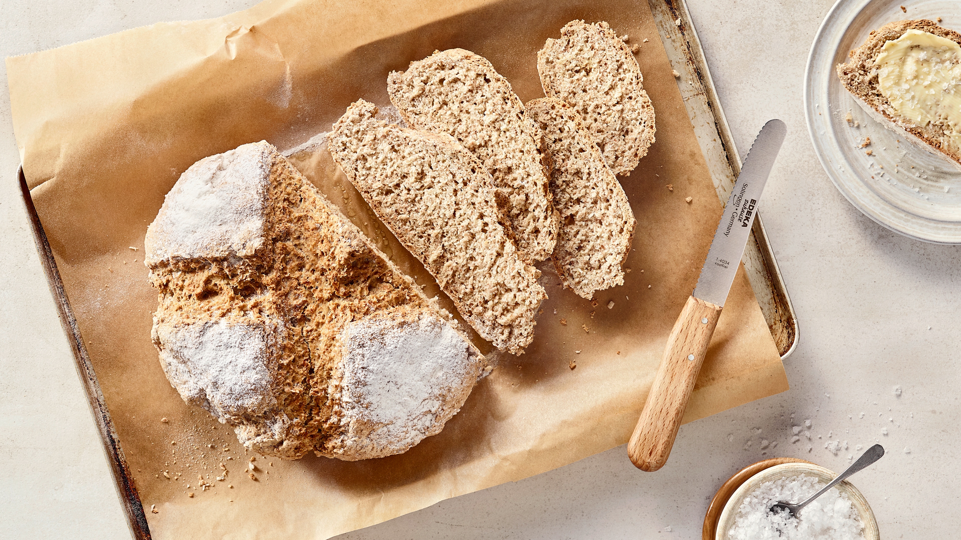 Ein Blech mit einem runden, aufgeschnittenen Brotlaib, mehreren hellbraunen Brotscheiben und weißem Mehl auf der Kruste.