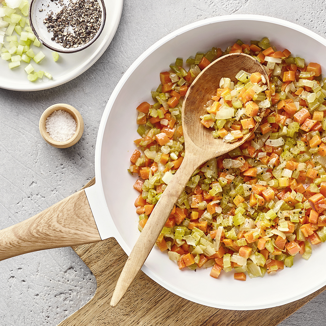 Tiefe, weiße Pfanne mit fein gewürfelten Karotten, Sellerie und Zwiebeln auf einem Holzbrett. Daneben stehen verschiedene Gewürze sowie ein Teller mit zusätzlichem Gemüse.