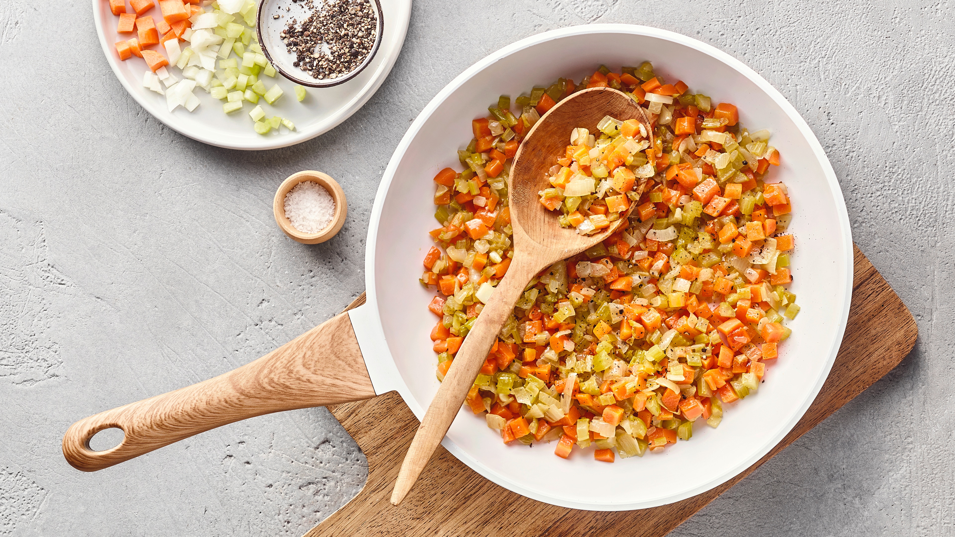 Tiefe, weiße Pfanne mit fein gewürfelten Karotten, Sellerie und Zwiebeln auf einem Holzbrett. Daneben stehen verschiedene Gewürze sowie ein Teller mit zusätzlichem Gemüse.