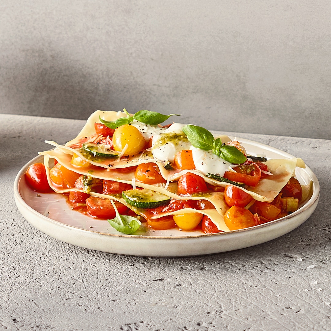 Ein Teller mit geschichteten Nudelplatten, dazwischen Tomaten, Zucchini & grünes Pesto, garniert mit heller Sauce & Kräutern