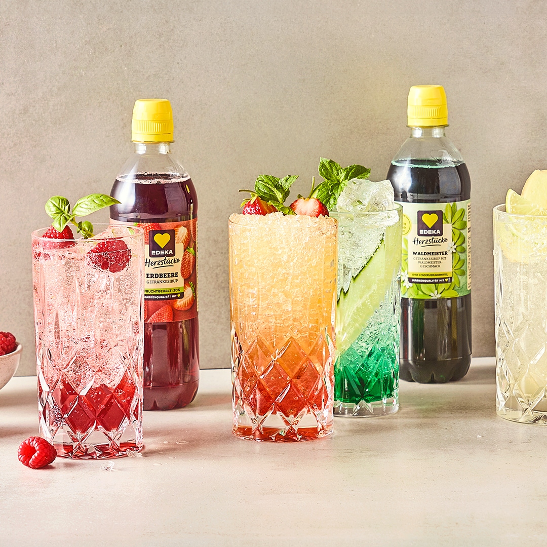 Cocktailgläser, unterschiedlich befüllt mit einem roten, orangem, grünen und gelben Getränk, daneben EDEKA Herzstücke Sirupe