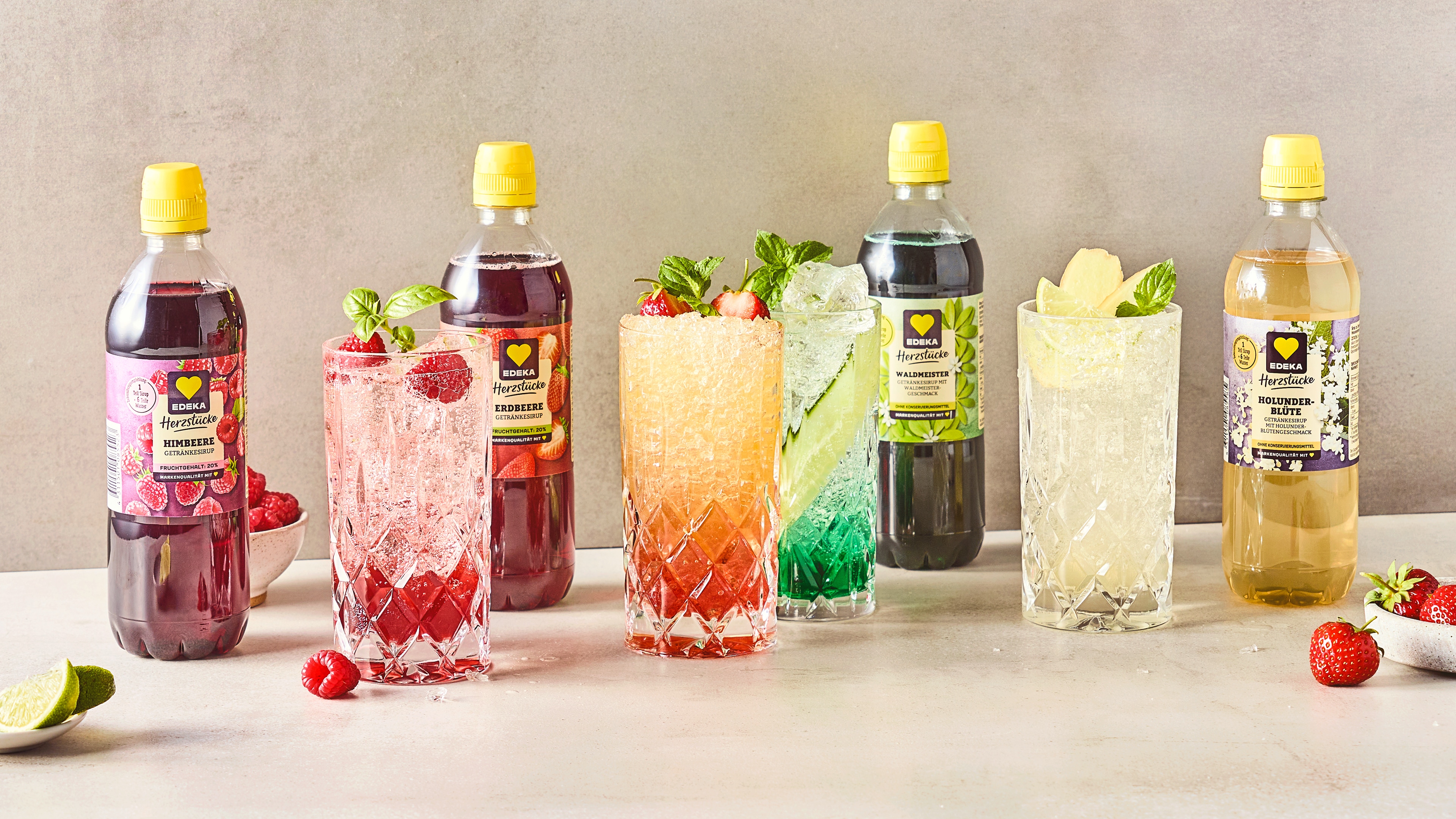 Rezept für Sommercocktails mit EDEKA Herzstücke Sirupen Cocktailgläser, unterschiedlich befüllt mit einem roten, orangem, grünen und gelben Getränk, daneben EDEKA Herzstücke Sirupe