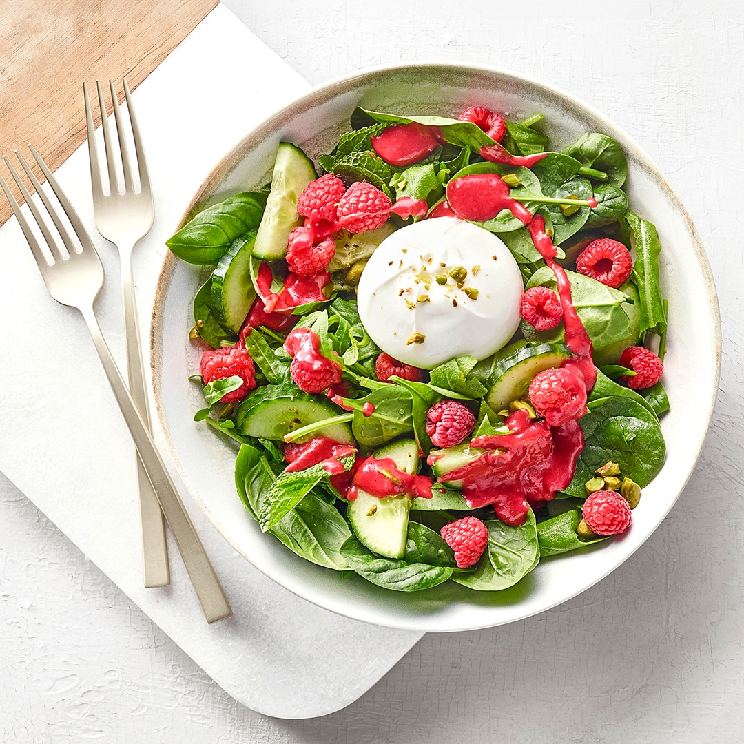 Salatteller mit Salatblättern, Gurke, Himbeeren, einem rosafarbenem Dressing und einem Klecks weißem Dressing. Links daneben zwei Gabeln.