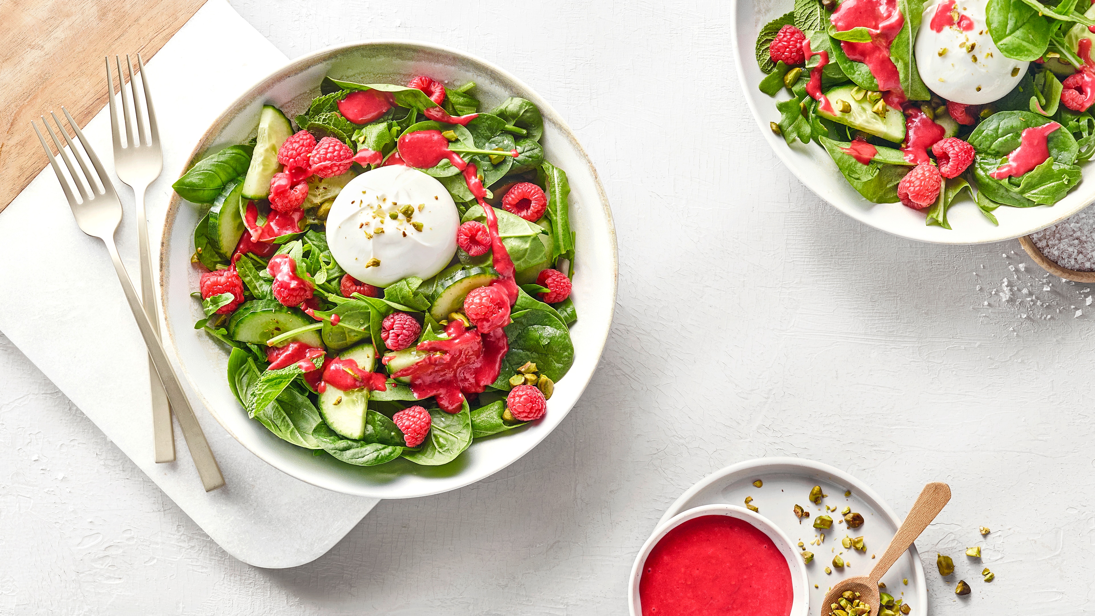 Salatteller mit Salatblättern, Gurke, Himbeeren, einem rosafarbenem Dressing und einem Klecks weißem Dressing. Links daneben zwei Gabeln. Oben rechts ein weiterer Teller mit Salat.