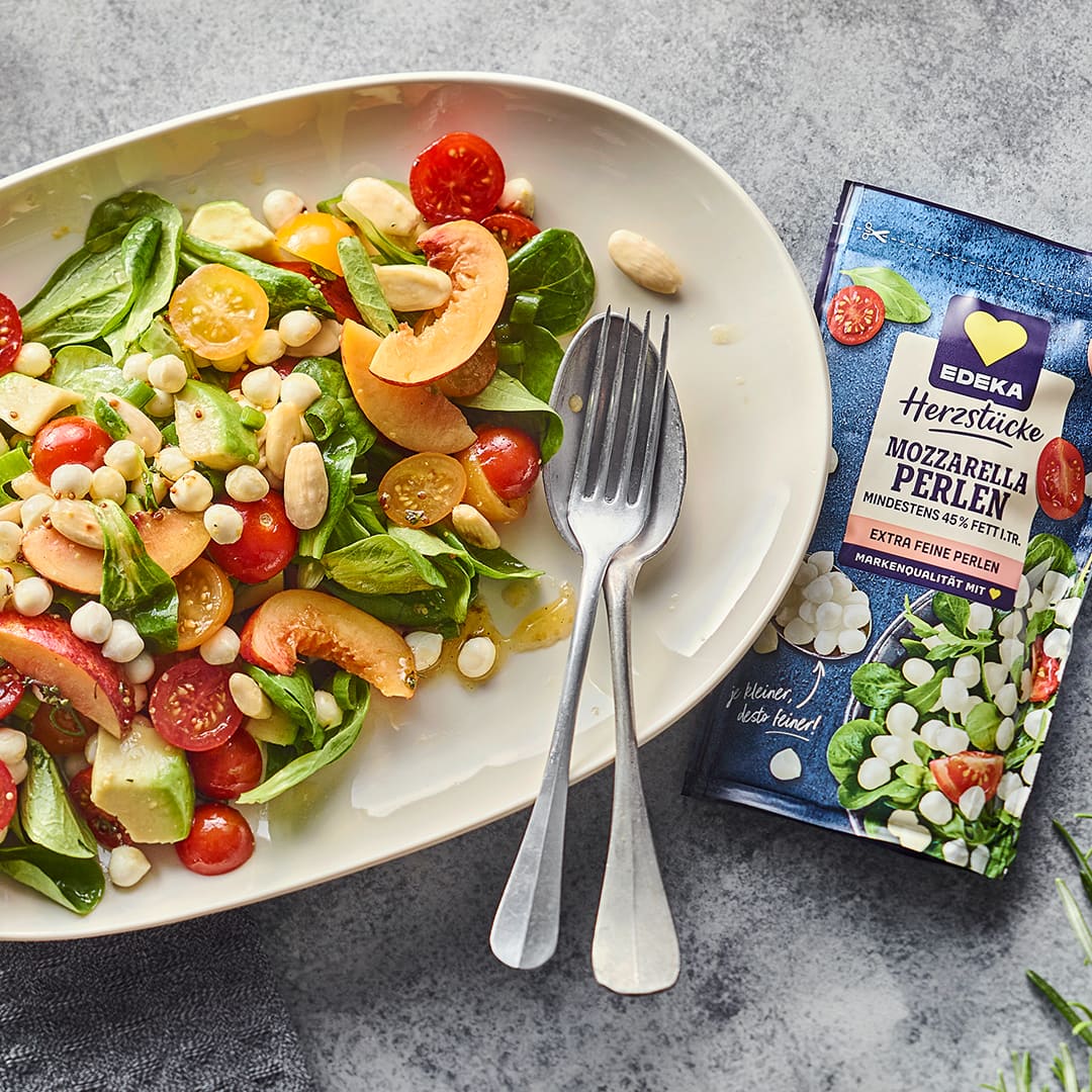 Ovaler Teller mit Salat mit Tomaten, Pfirsich und Mozzarella Perlen, daneben die EDEKA Herzstücke Mozzarella Perlen Packung.