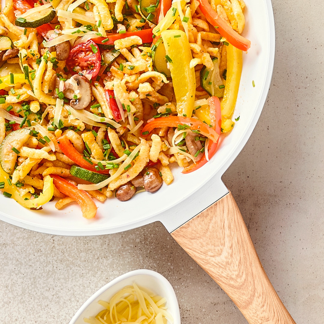 Eine Pfanne mit Spätzle, Paprikastreifen, Kirschtomaten, Champignons und Zucchini,  getoppt mit Schnittlauch und Reibekäse