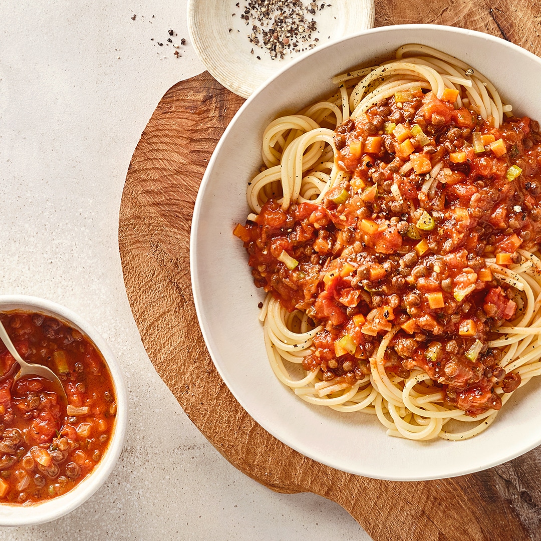 Ein Teller mit Spaghetti, darauf rote Soße mit kleinen Würfeln von Gemüse und braunen Linsen, daneben eine Schale mit Soße