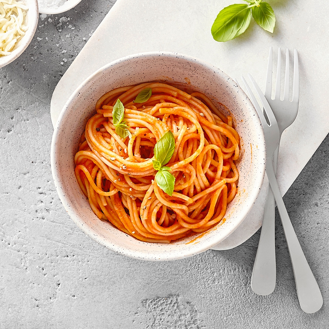 Eine Schale Spaghetti mit Tomatensauce und Basilikum, dazu geriebener Käse und Besteck.