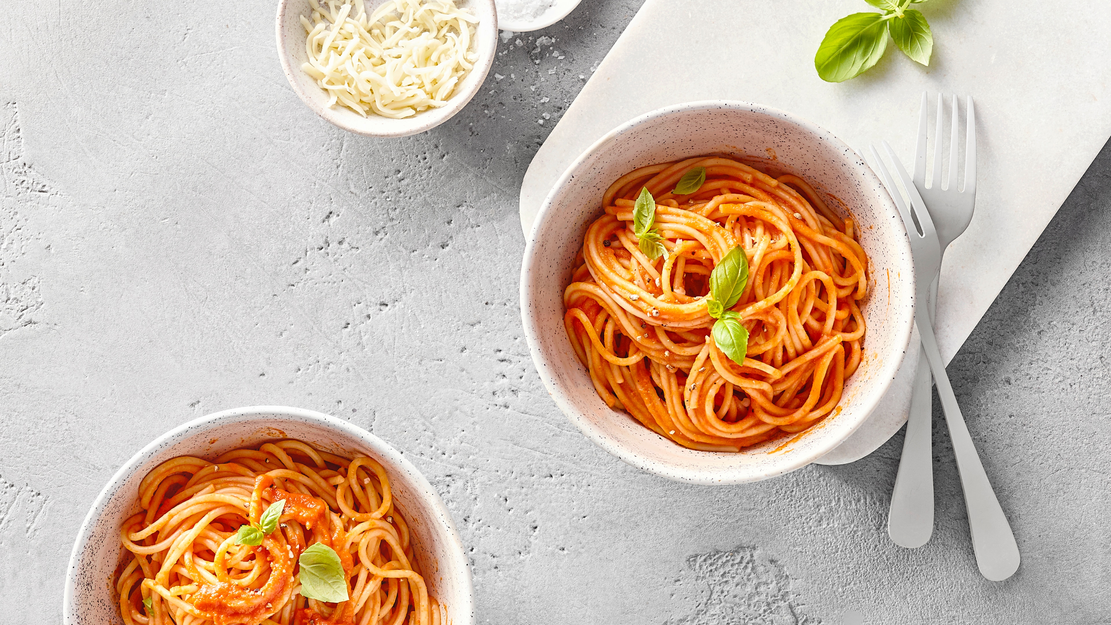 Zwei Schalen Spaghetti mit Tomatensauce und Basilikum, dazu geriebener Käse und Besteck.