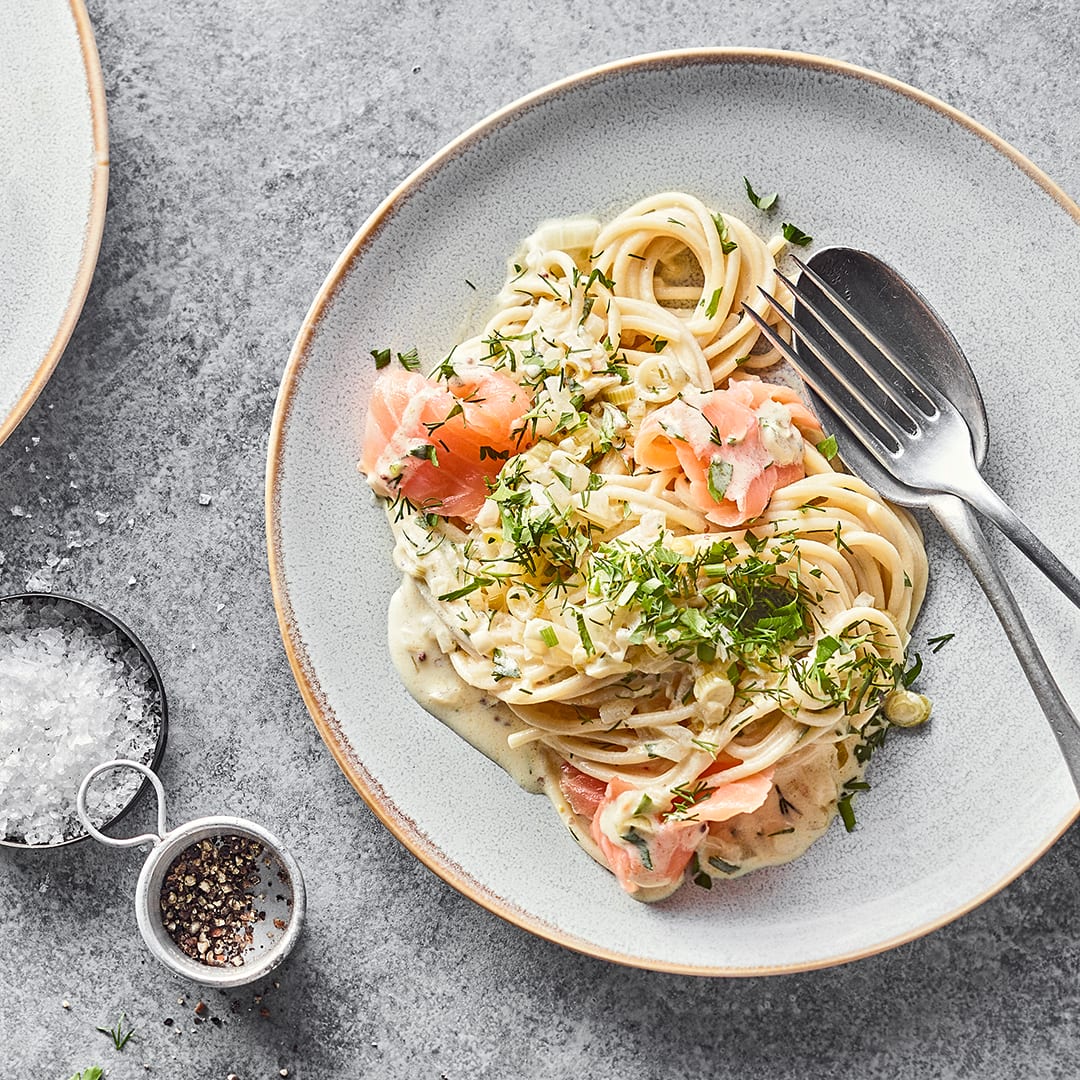 Ein runder Teller mit cremigen Zitronen-Spaghetti mit Räucherlachs, getoppt mit frischem Dill und Petersilie