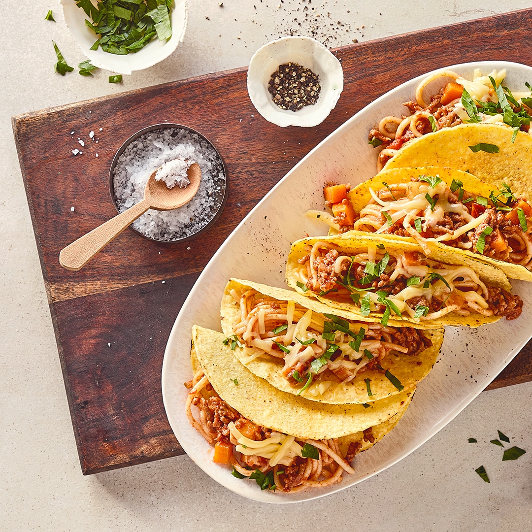 Ein ovaler Teller mit Tacoschalen, gefüllt mit Spaghetti und Hackfleisch, getoppt mit Streukäse und gehacktem Koriander